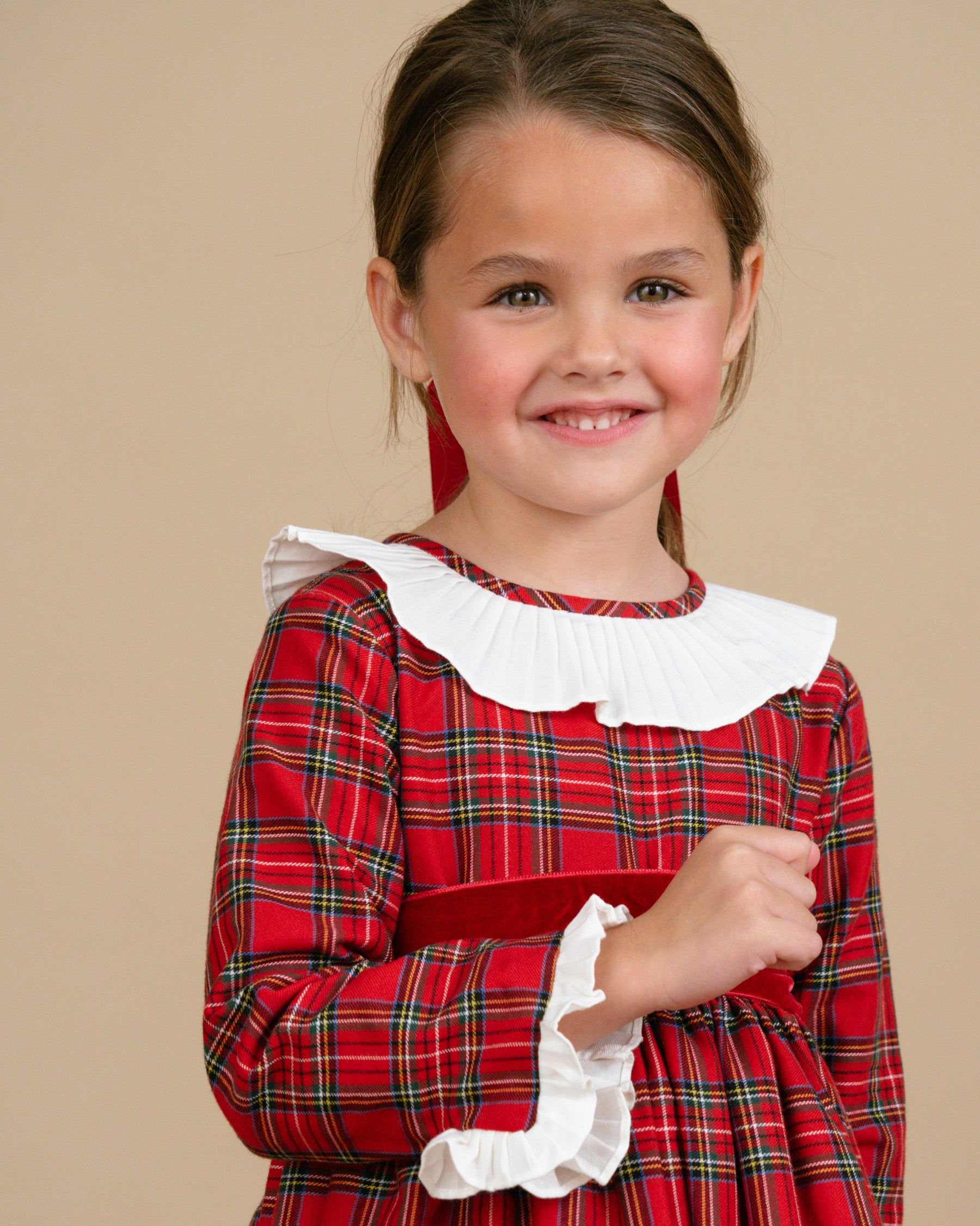 Young girl wearing a red plaid dress with white ruffled collar against a beige background