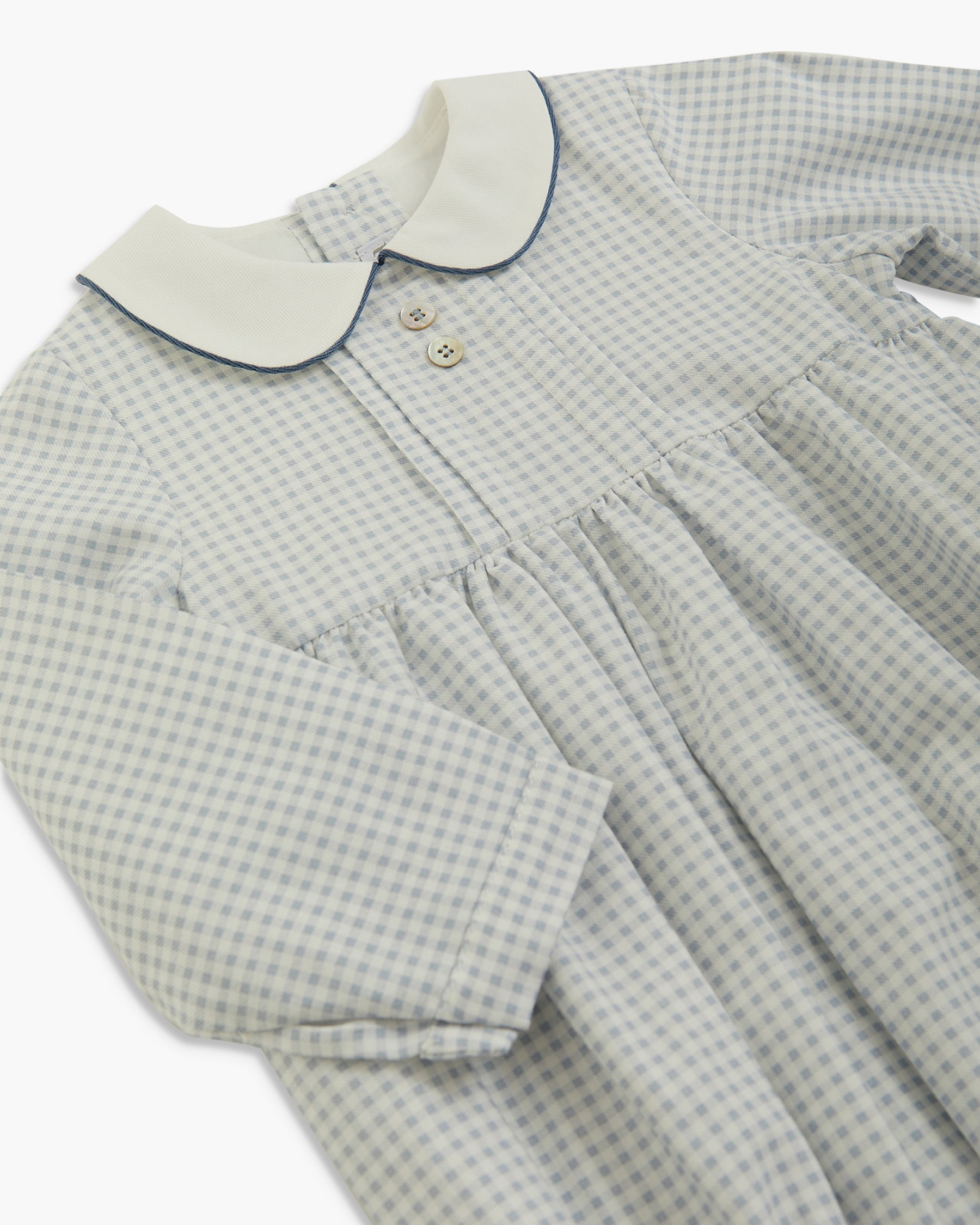 A close-up of a long-sleeve baby dress with a light gray and white gingham pattern, featuring a contrasting cream-colored collar with dark piping and two small buttons.