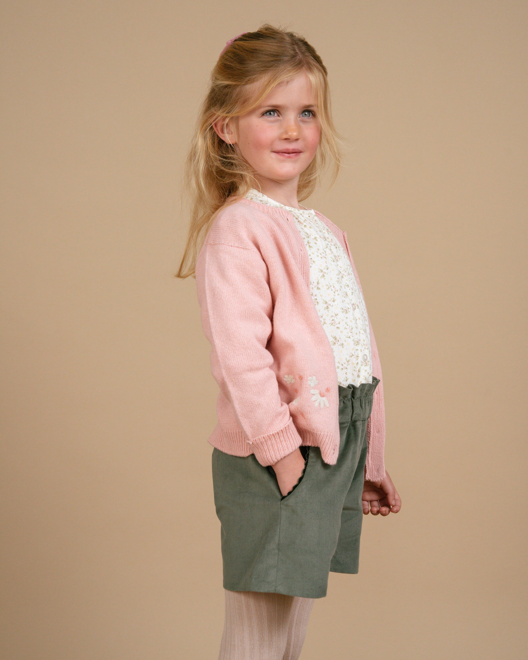 Young girl wearing a pink cardigan with lace details and green shorts on a beige background