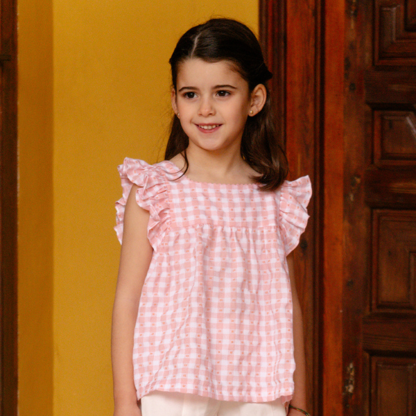 Young girl wearing a pink checkered top standing in front of a wooden door.