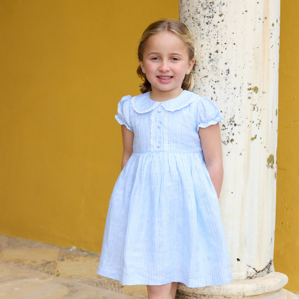 Young girl in a light blue dress standing against a yellow wall.