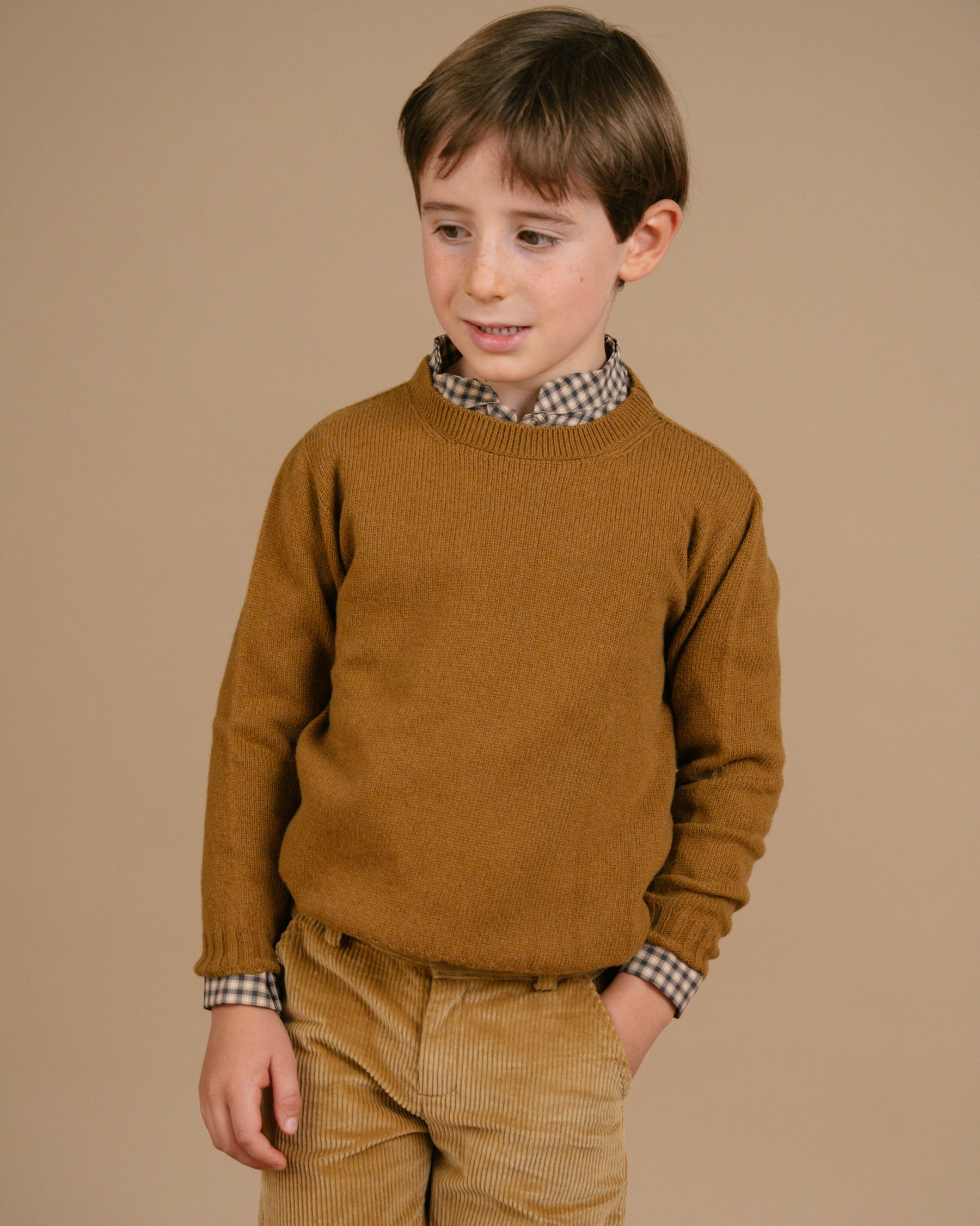 Young boy wearing a mustard yellow sweater over a checkered shirt with matching pants on a brown background