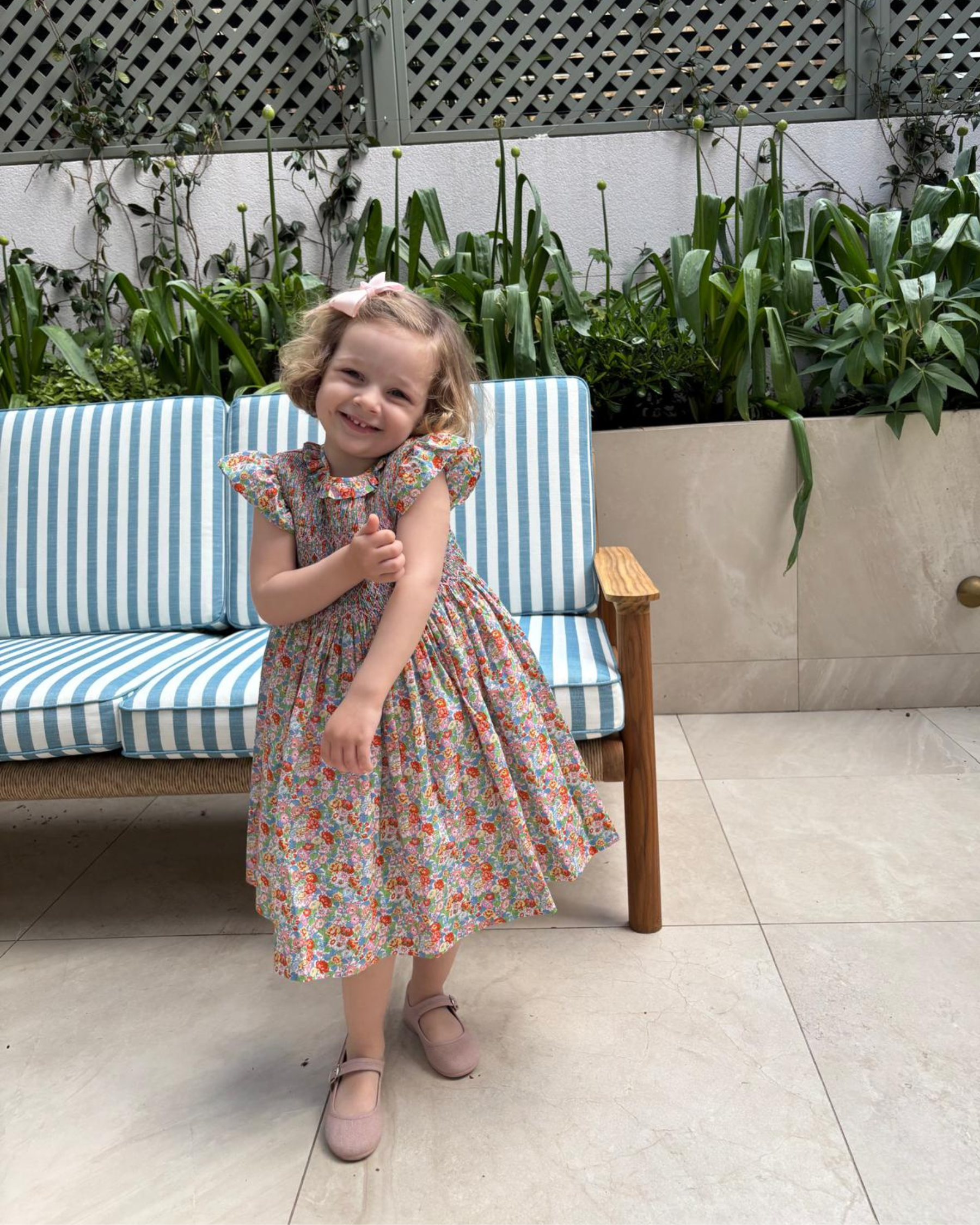 Young girl in a floral dress standing next to a striped bench with plants in the background