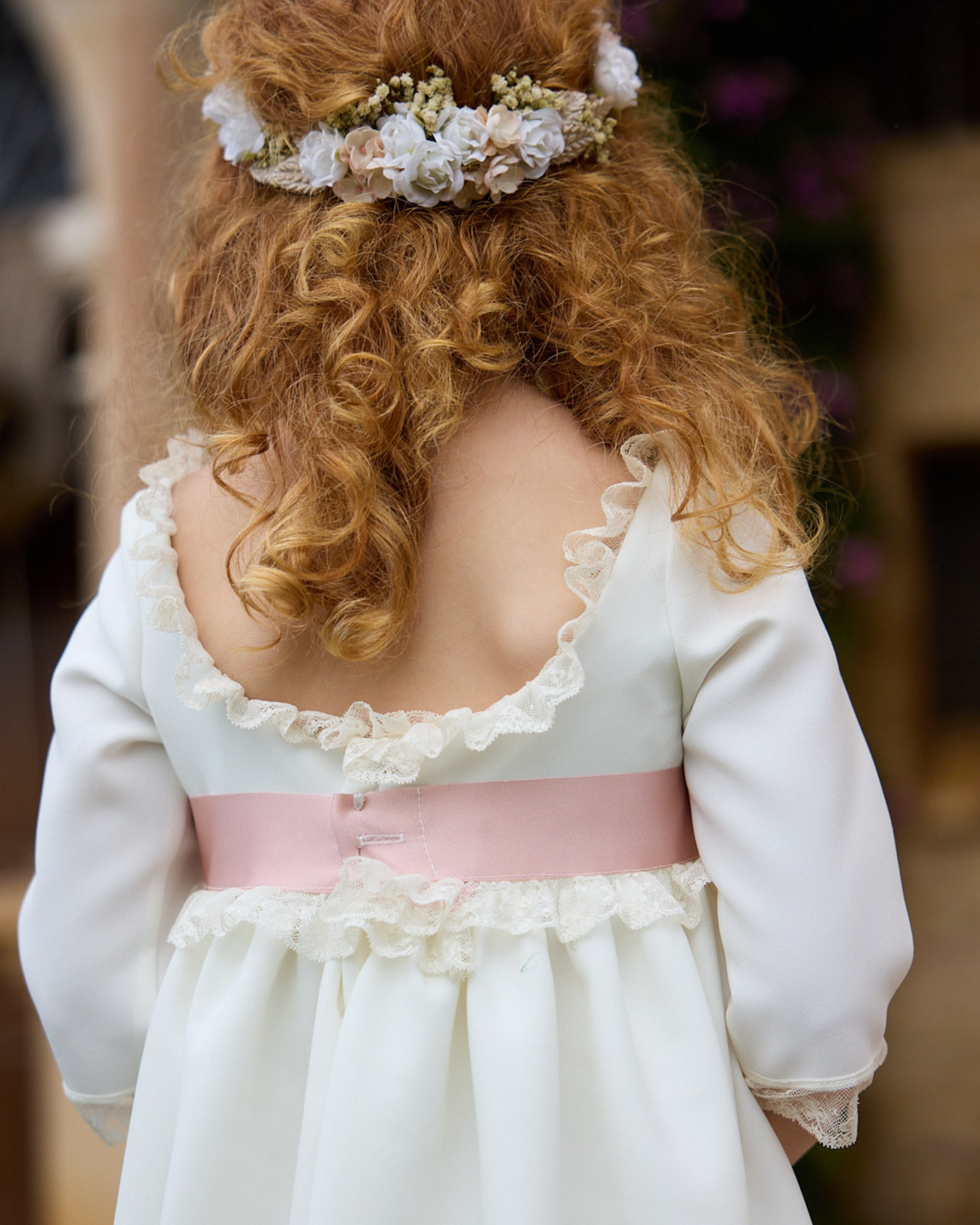 Child wearing a white dress with a pink sash and floral headband in a blurred indoor setting