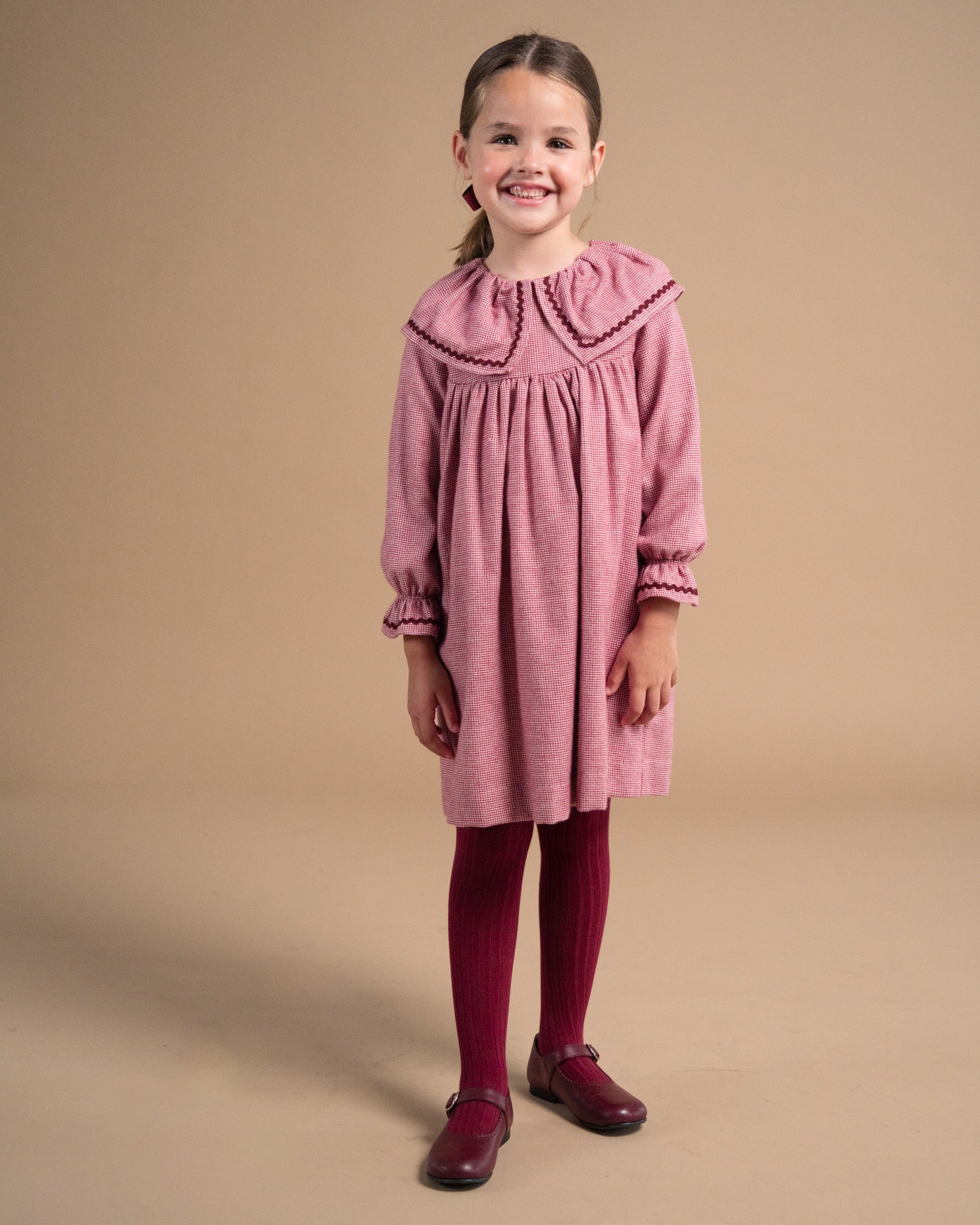 A young girl stands against a solid beige studio background wearing a dusty rose pink long-sleeve dress with a wide embroidered collar, dark red tights, and dark red shoes.