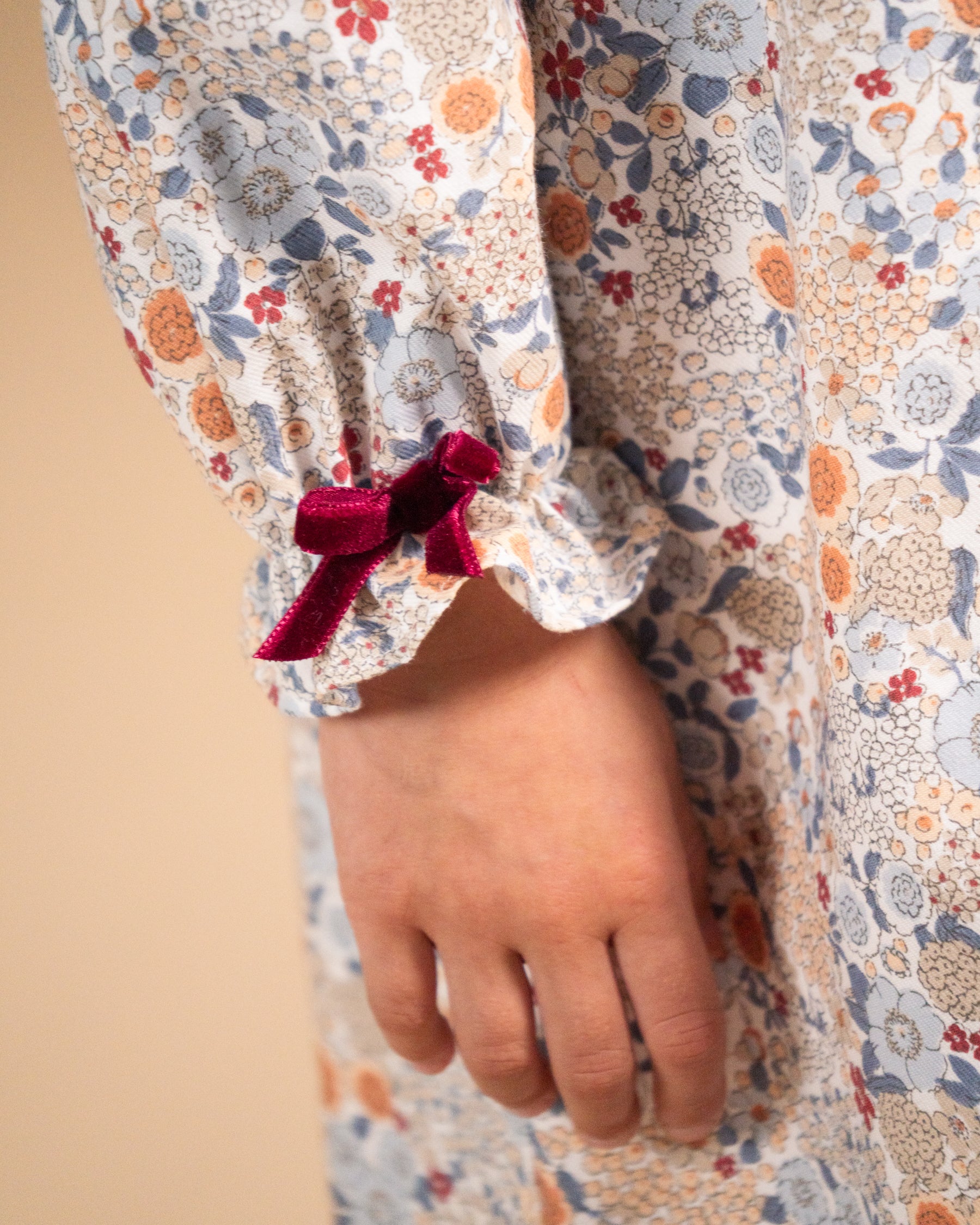 Close-up of a child wearing a long-sleeve floral dress with a gathered cuff and a dark red velvet bow, against a plain beige background.