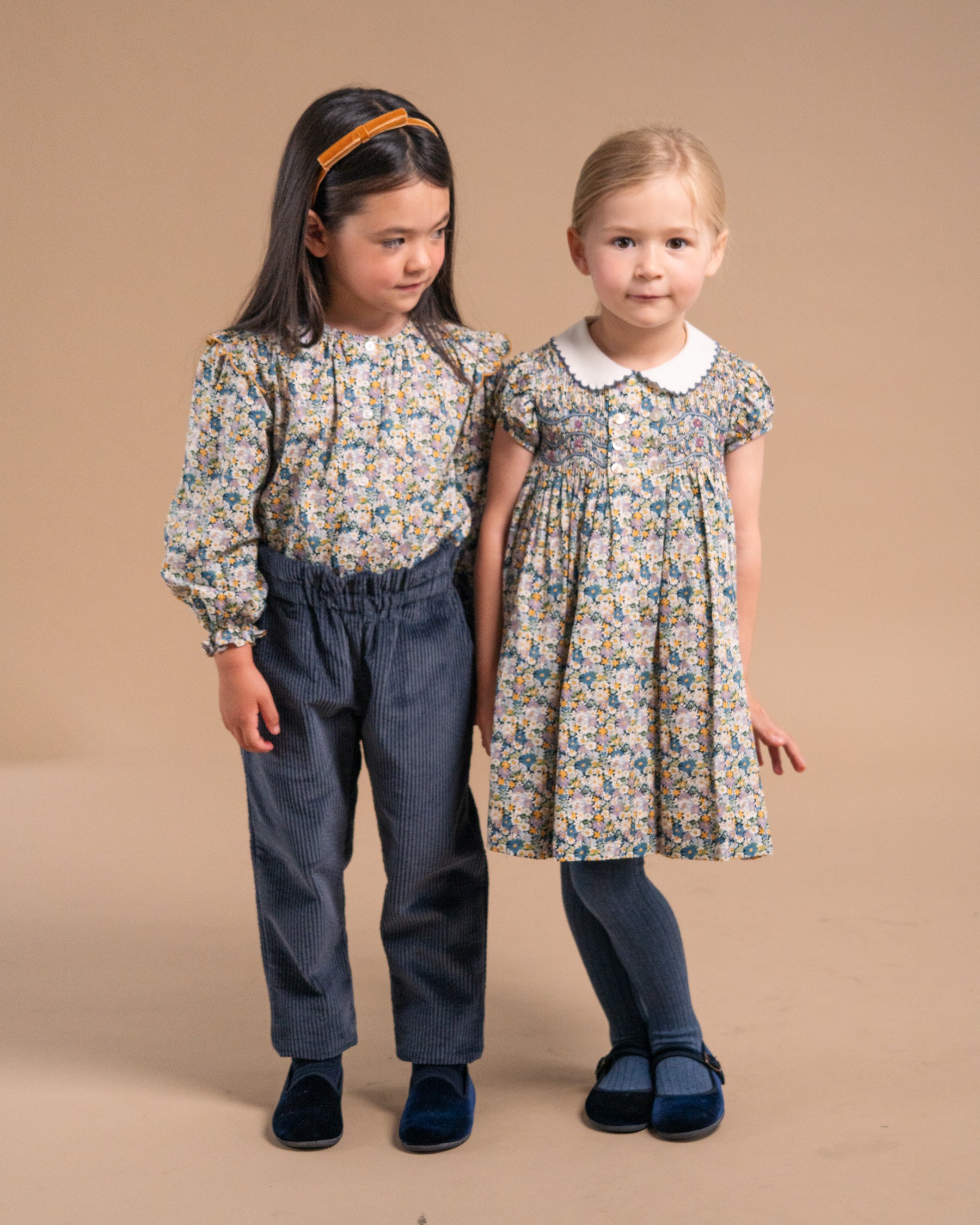 Two young girls modeling matching floral print clothing against a solid beige studio background. The girl on the left wears a long-sleeve floral top with dark blue corduroy pants, and the girl on the right wears a matching floral dress with a white Peter Pan collar and dark blue tights.