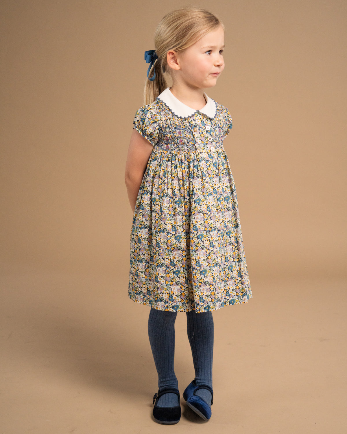 A young girl with blonde hair tied back with a blue bow is wearing a short-sleeved floral print dress with a white Peter Pan collar, blue tights, and blue shoes, standing against a solid beige studio background.