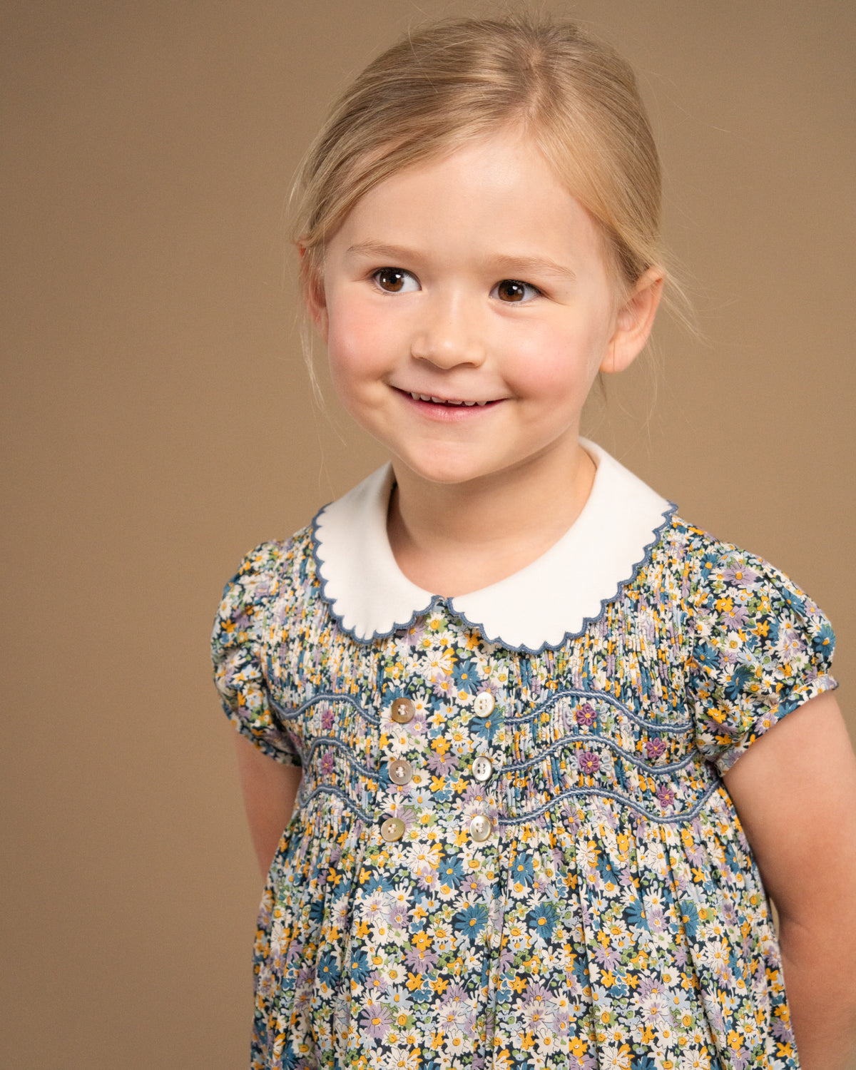 A young girl with blonde hair smiling, wearing a short-sleeve floral dress with a white Peter Pan collar, photographed against a solid warm brown studio background.