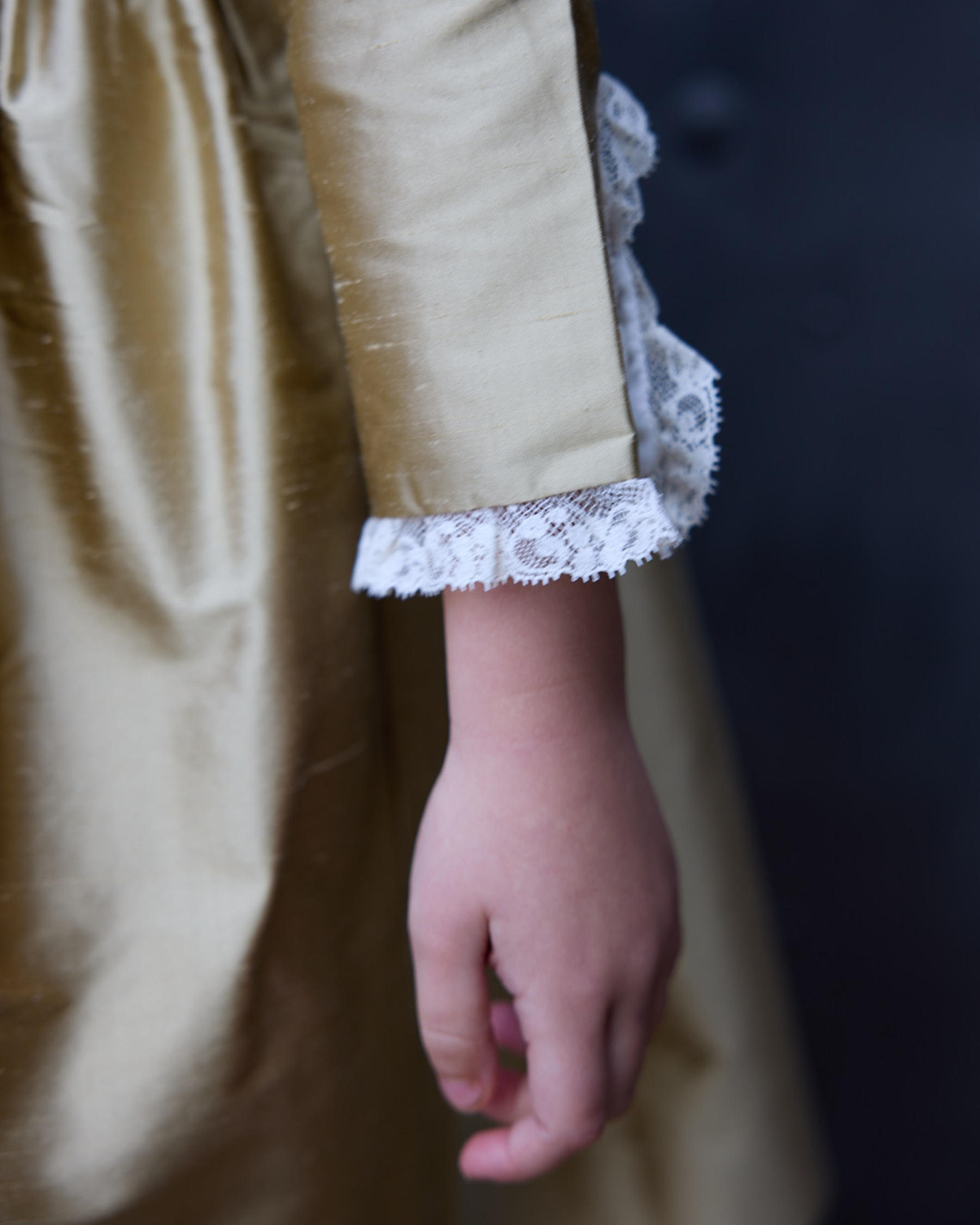 Close-up of a child's hand wearing a beige dress with lace cuffs against a dark background