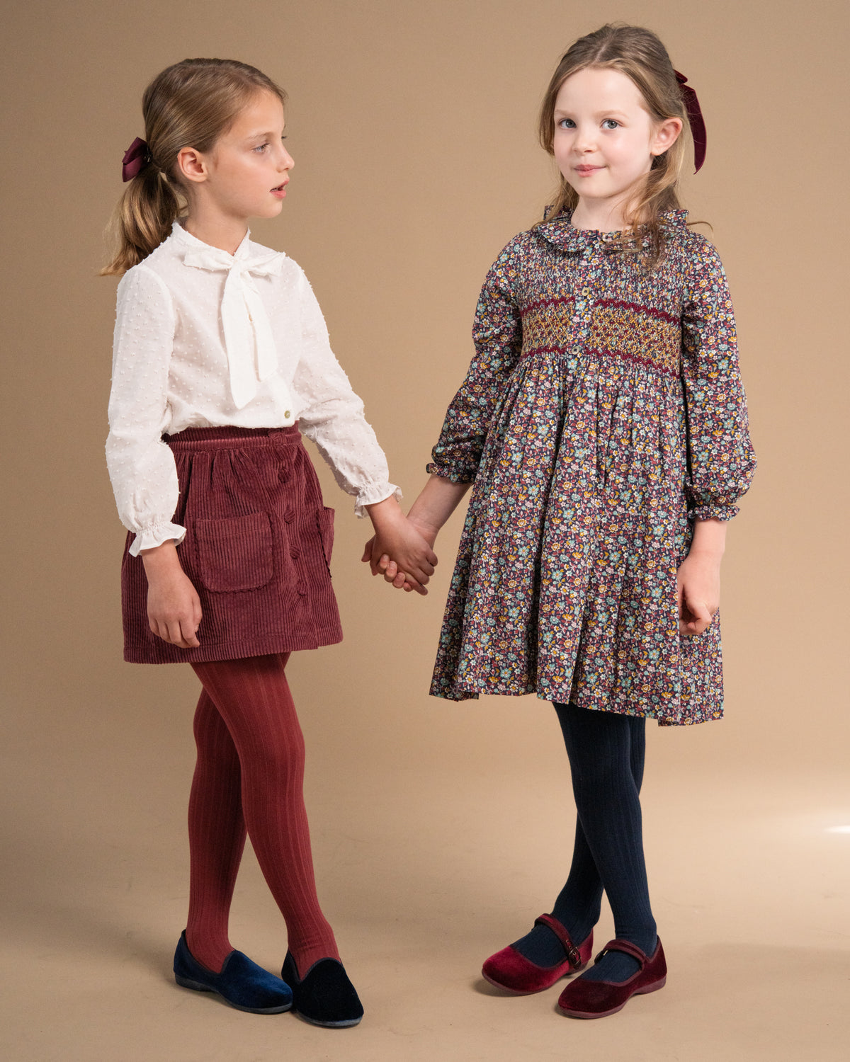 Two young girls holding hands in a studio setting, wearing coordinated outfits. The girl on the left wears a white blouse, burgundy skirt, burgundy tights, and dark blue shoes.