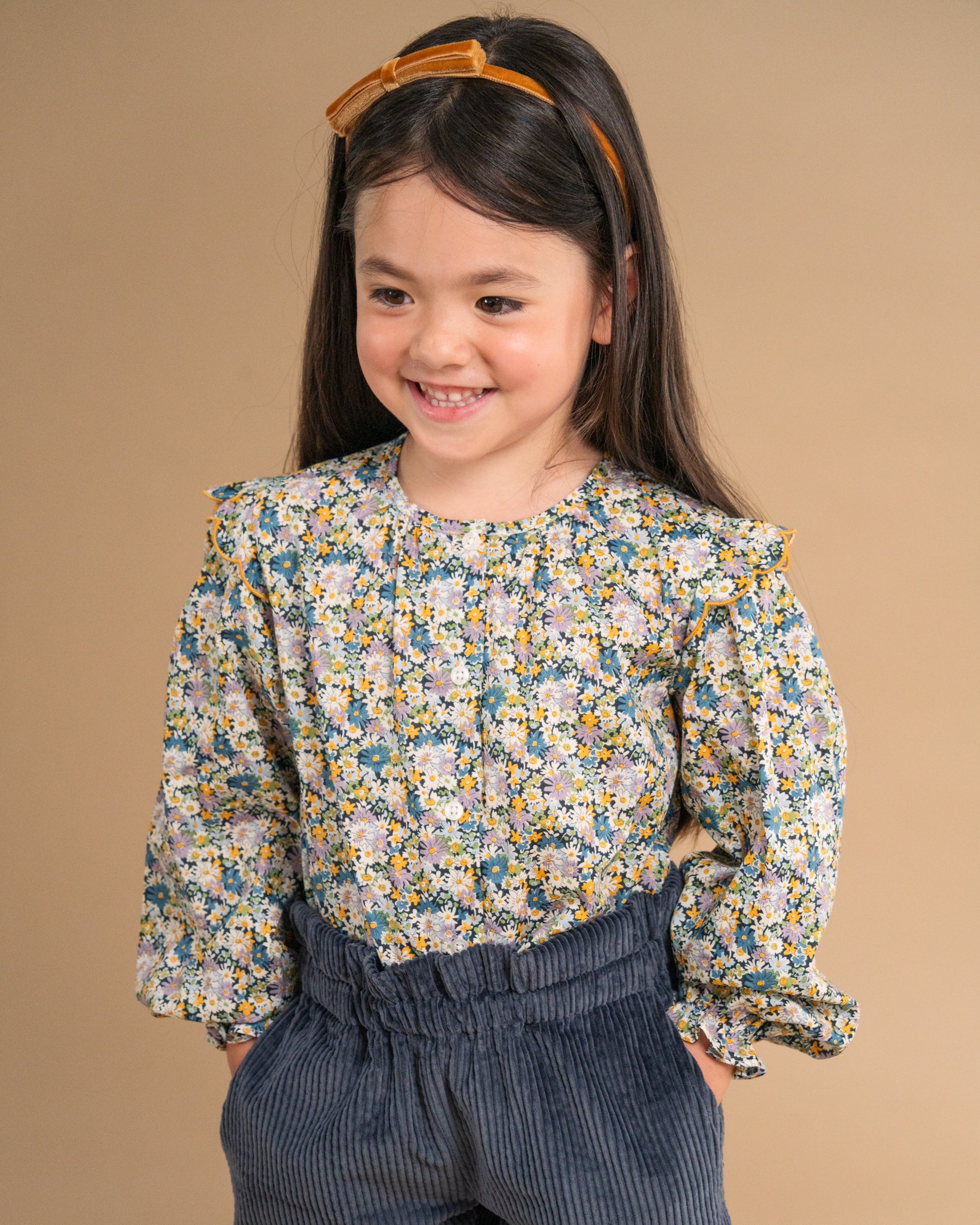 A young girl smiling and looking to the side, wearing a long-sleeve floral blouse with a blue, yellow, and white pattern, paired with dark blue corduroy pants, set against a solid beige background.