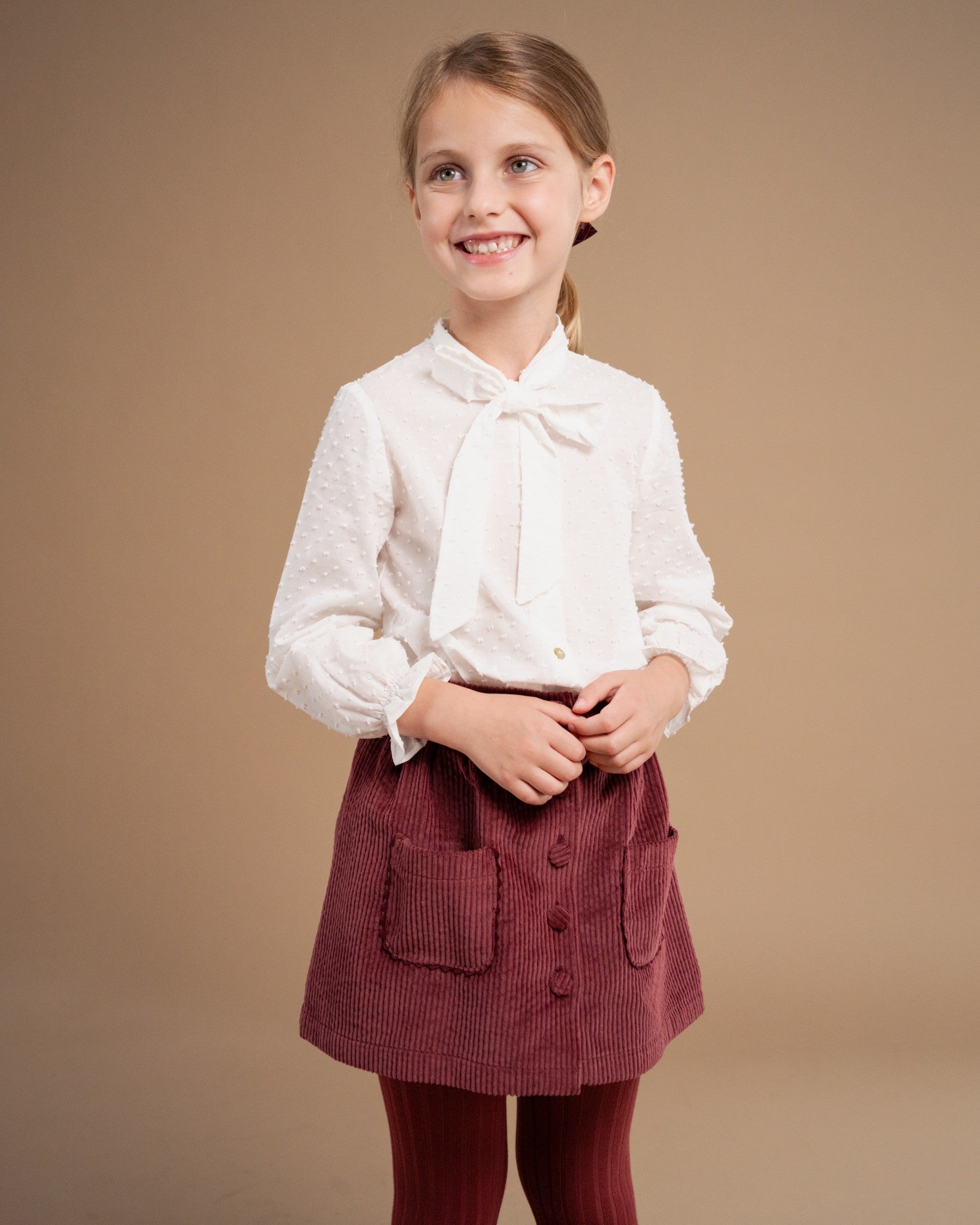 A young girl smiling and posing in a studio setting, wearing a white long-sleeve blouse with a bow collar and a burgundy corduroy skirt with front pockets.
