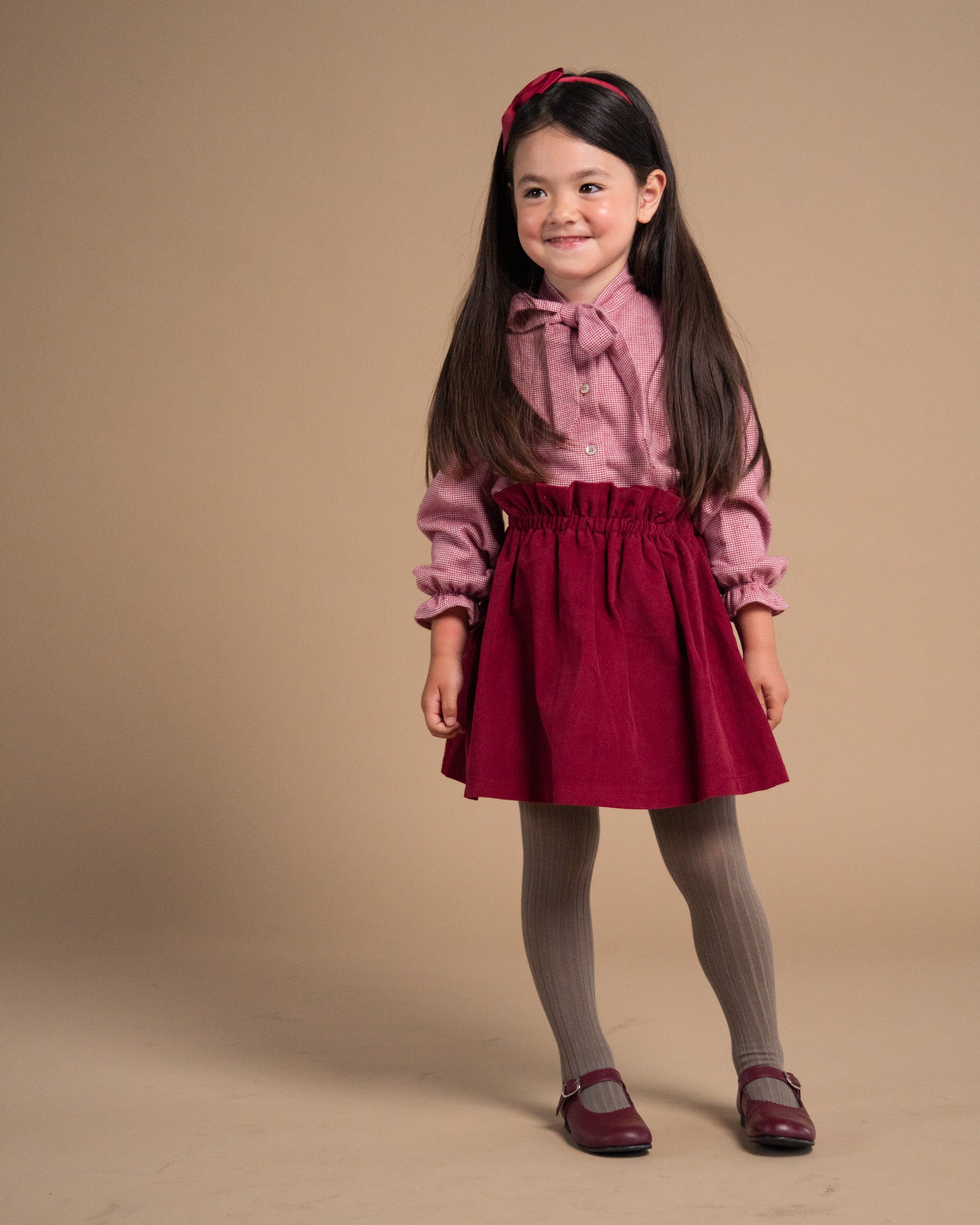 A young girl with long dark hair stands against a solid beige studio background, wearing a dusty pink long-sleeve blouse with a bow collar, a dark red corduroy pleated skirt, gray tights, and burgundy shoes.