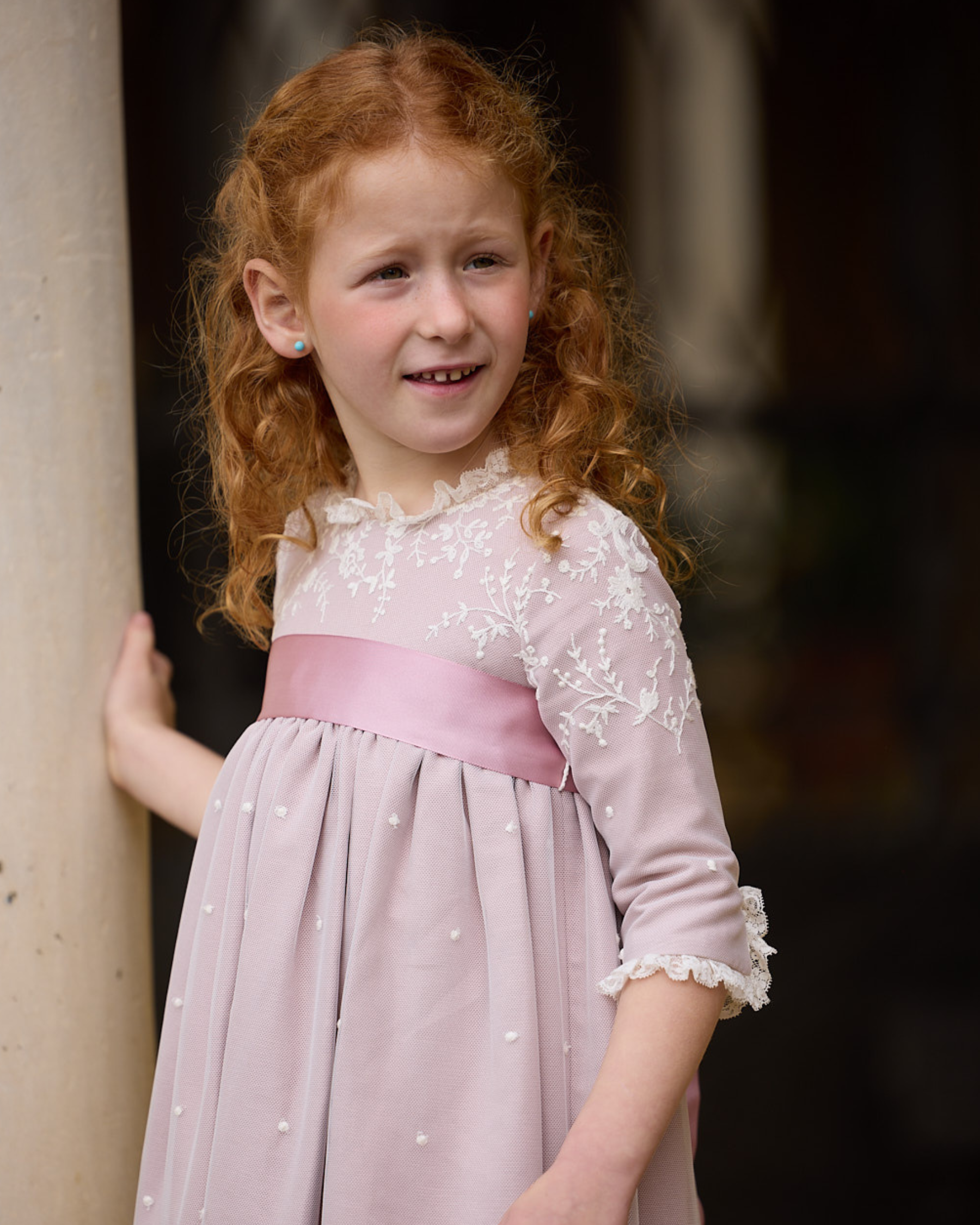 Young girl in a light purple dress with a pink ribbon standing against a blurred background