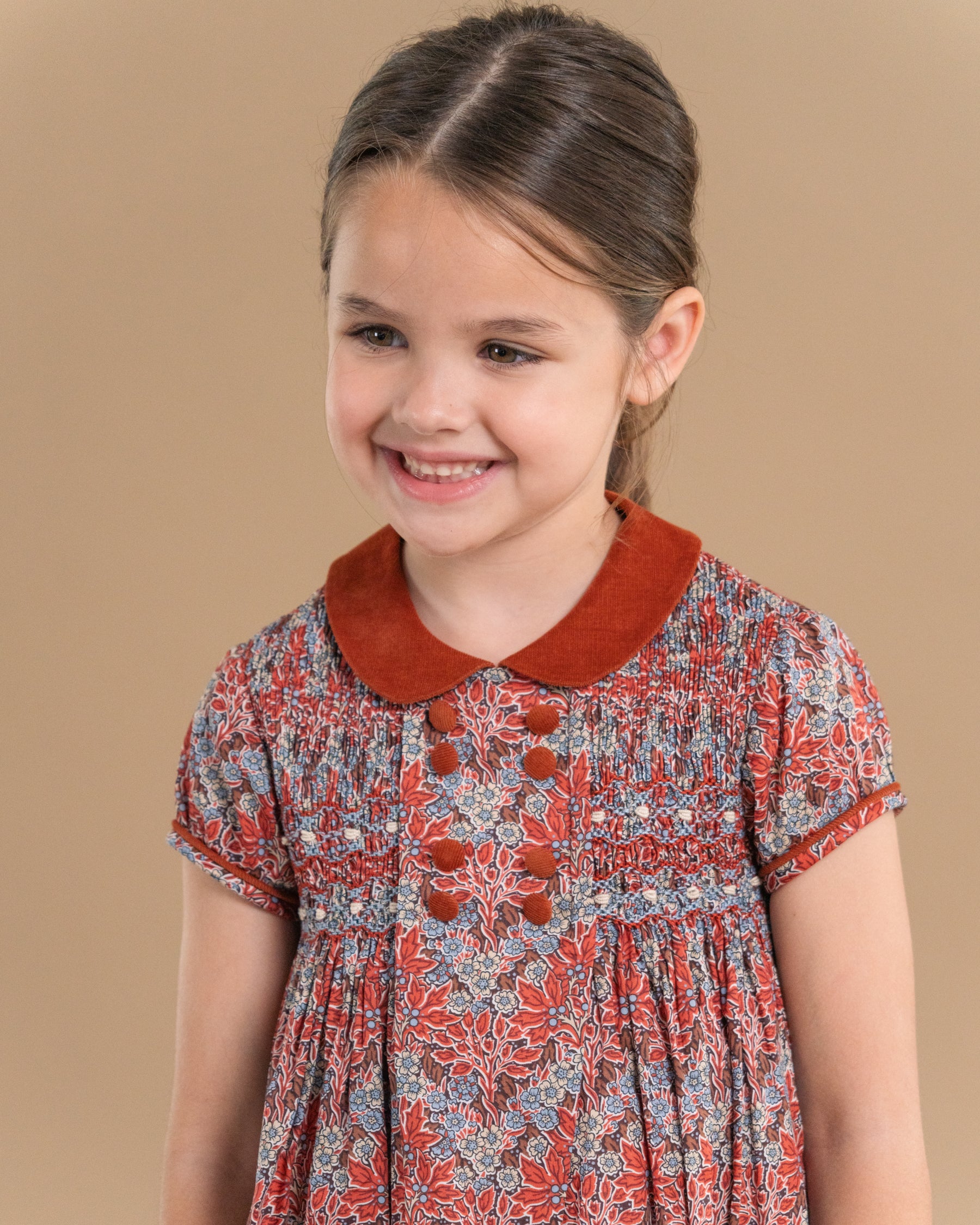 A young girl with brown hair pulled back, wearing a short-sleeve dress with a rust-colored collar and a red, blue, and white floral pattern, photographed against a solid tan background.