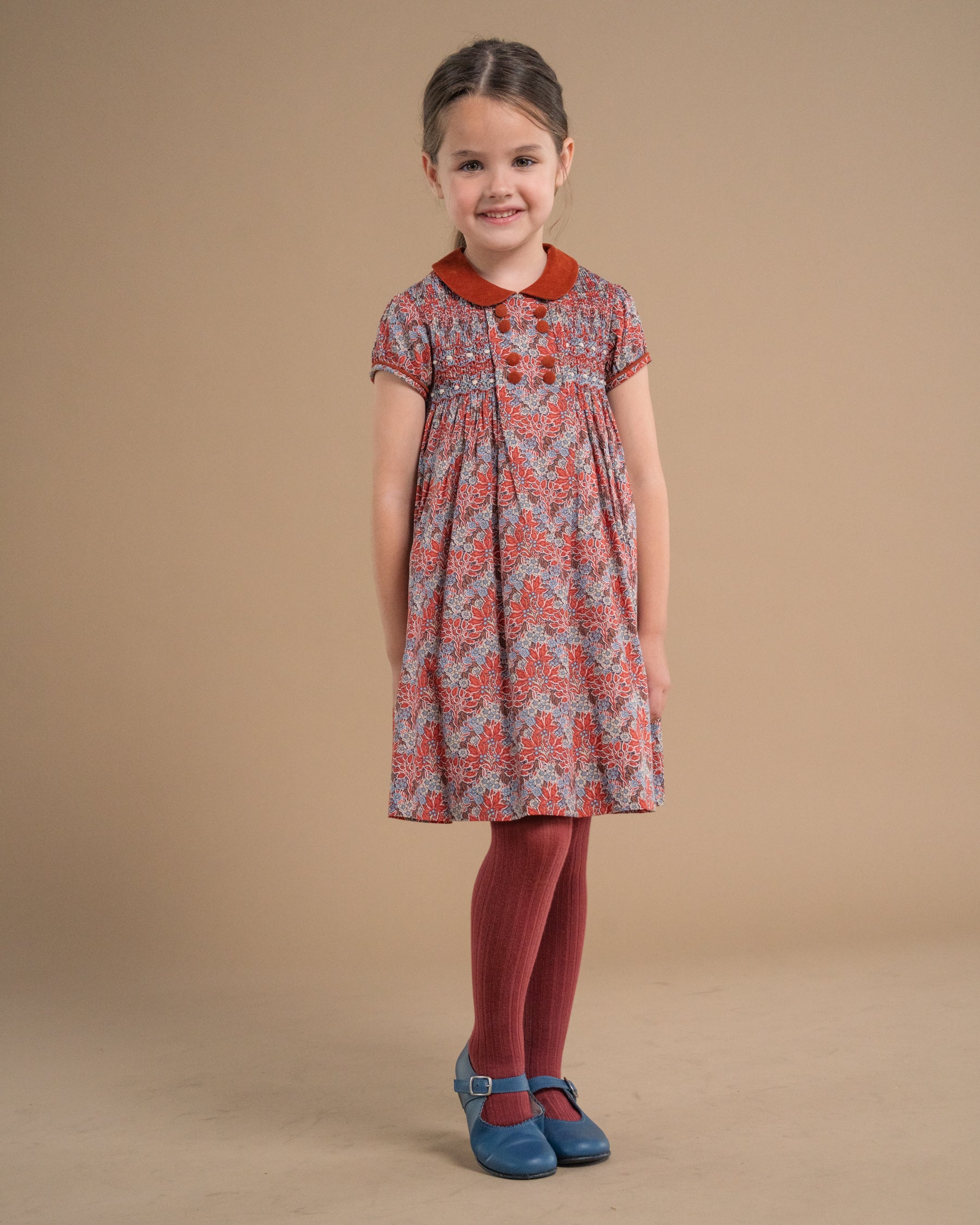 A young girl stands against a solid beige studio background wearing a short-sleeved dress with a red collar and a red, orange, and gray patterned body, paired with red tights and blue shoes.