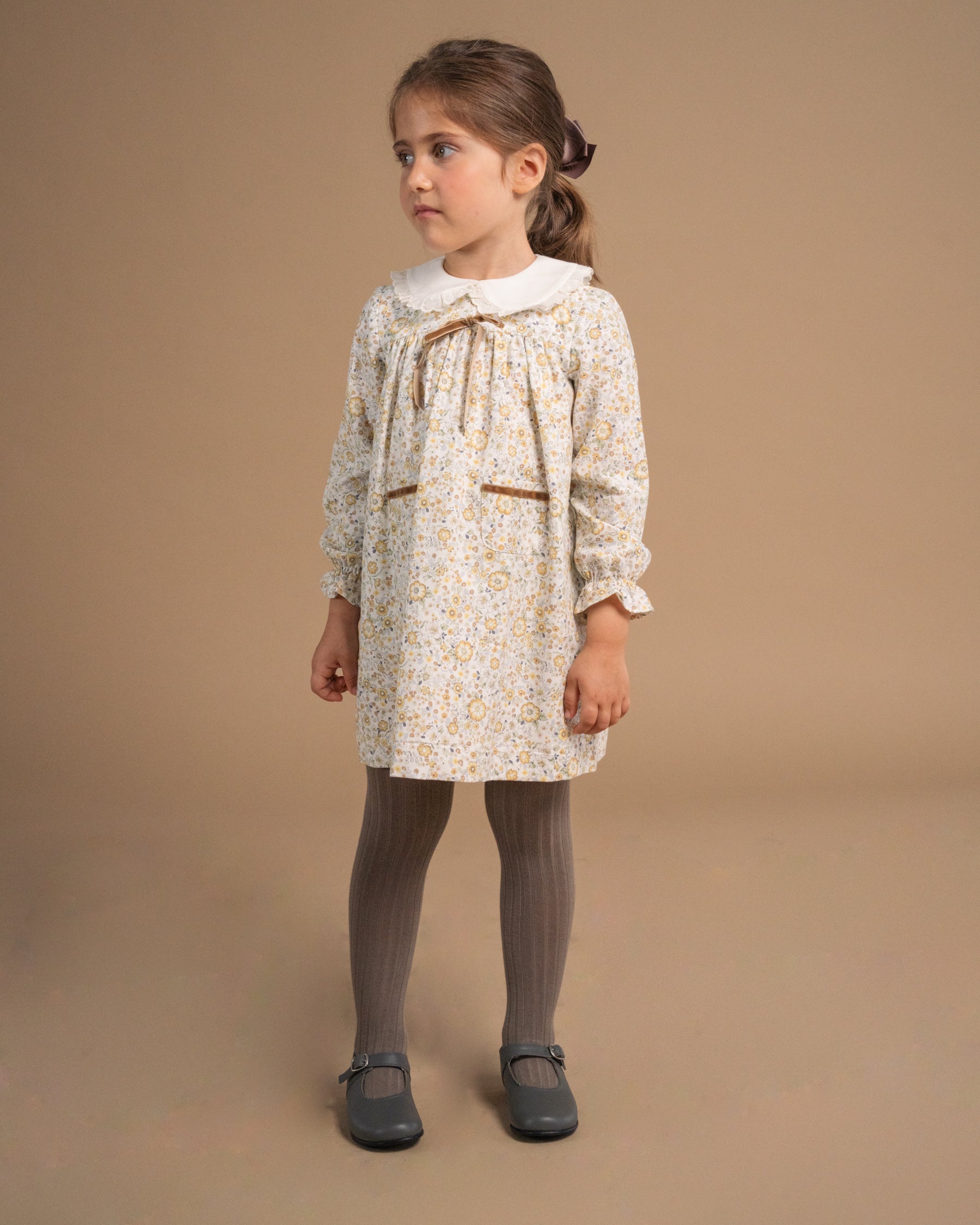 A young girl modeling a cream-colored floral dress with a white Peter Pan collar, paired with gray tights and dark gray shoes, standing against a solid beige studio background.