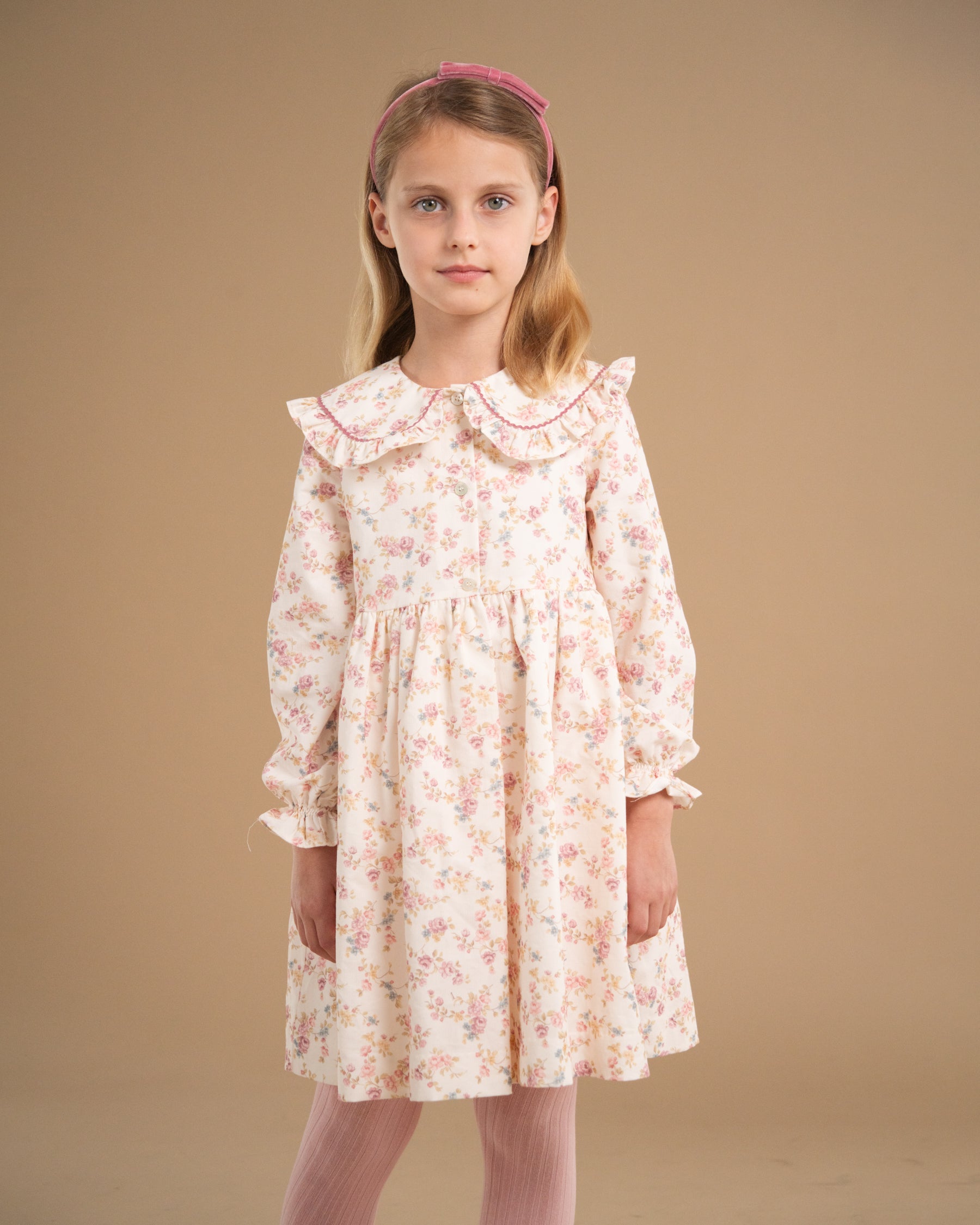 A young girl with blonde hair wearing a pink floral long-sleeve dress with a ruffled Peter Pan collar, standing against a solid beige studio background.