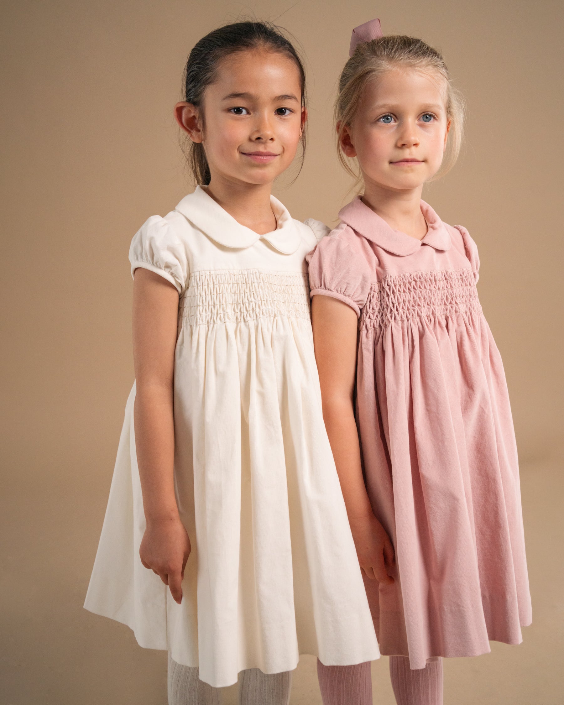 Two young girls modeling smocked dress dresses, one in off-white and the other in dusty pink, standing against a solid beige studio background.