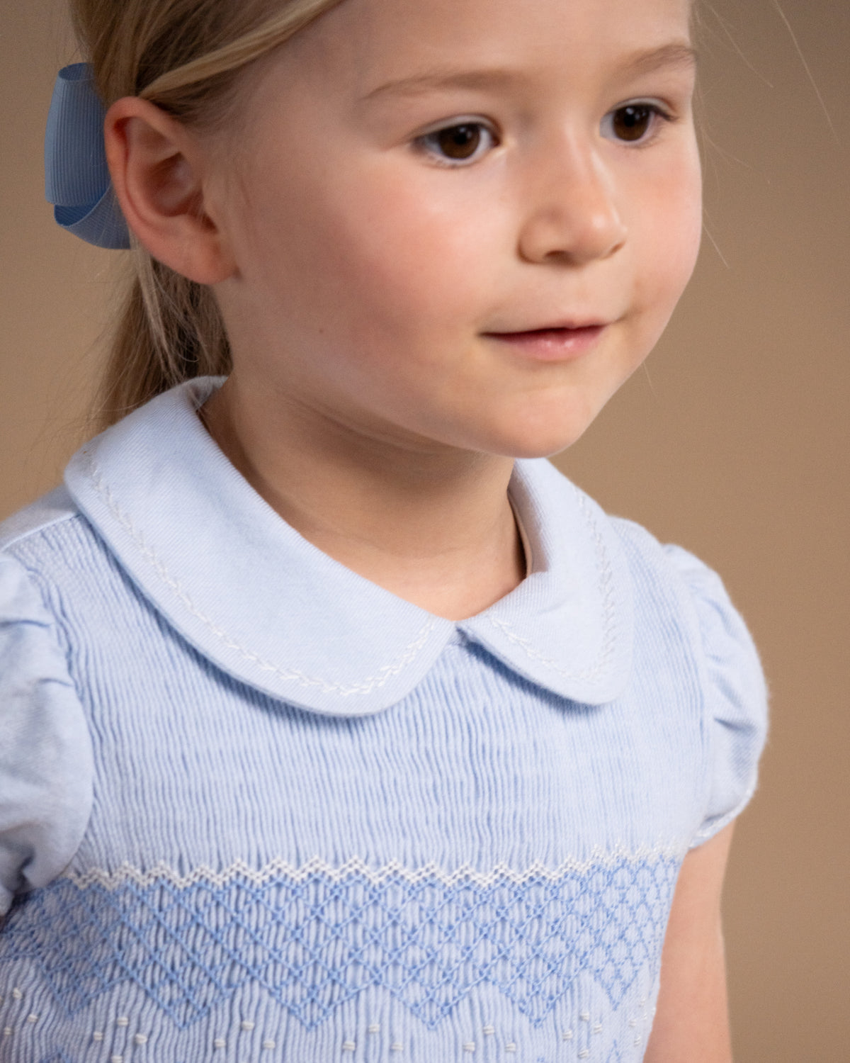 A young girl with blonde hair tied back with a blue bow, wearing a light blue smocked dress with a Peter Pan collar, shown in a close-up portrait against a warm beige background.