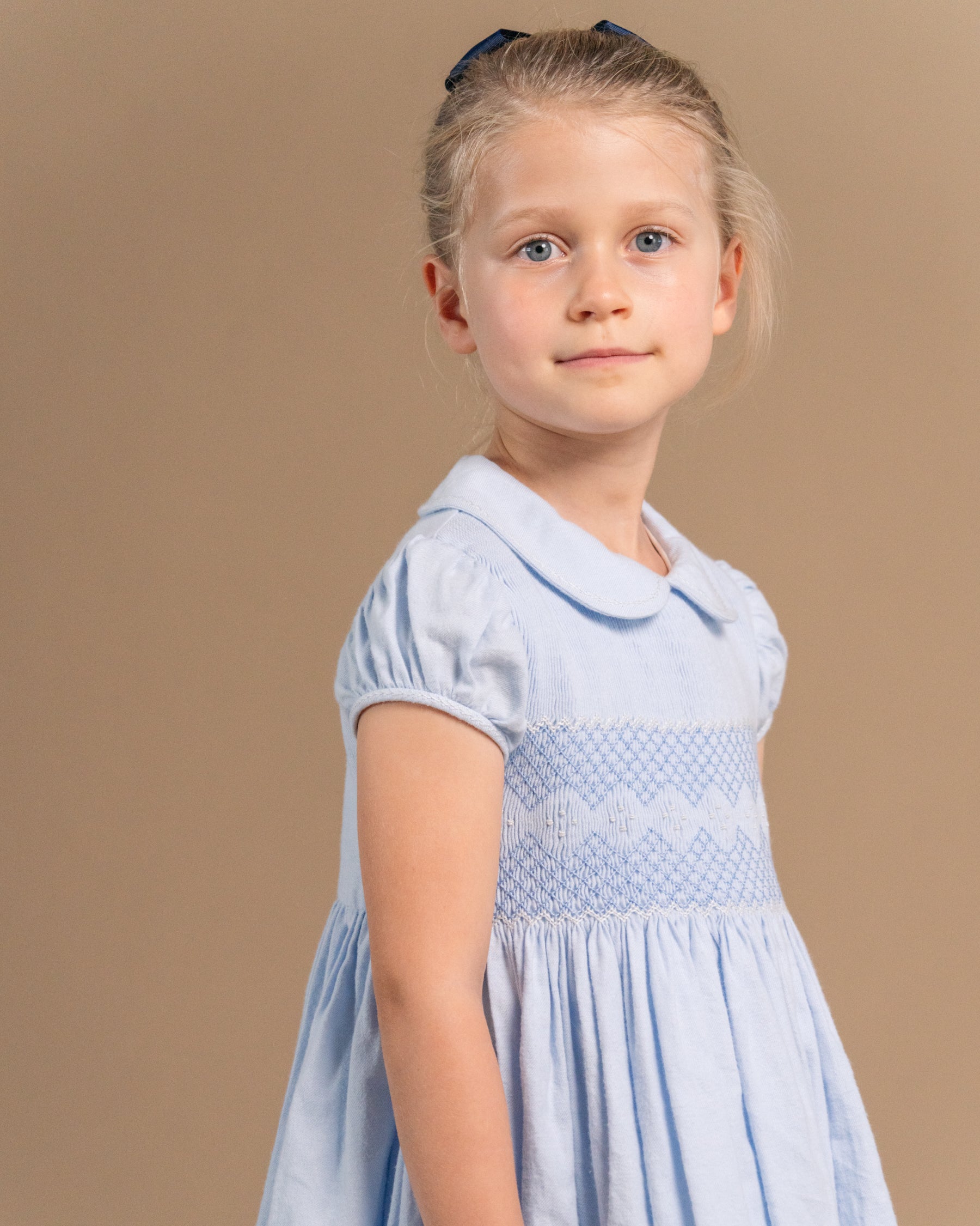 A young girl with blonde hair tied up with a dark bow is wearing a light blue short-sleeve dress with a Peter Pan collar and smocked bodice, photographed against a solid beige studio background.