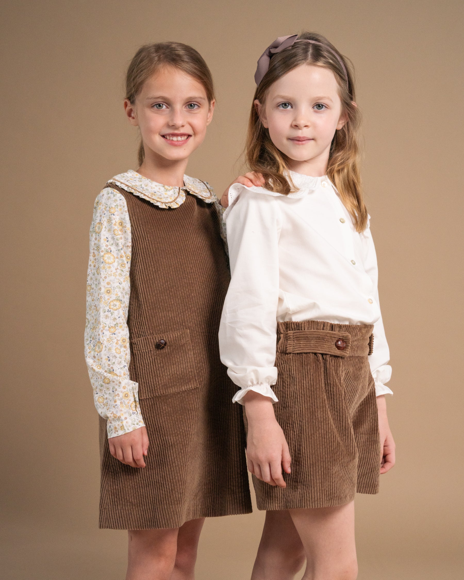 Two young girls modeling coordinated outfits against a solid warm beige studio background. The girl on the left wears a brown corduroy pinafore dress over a cream floral long-sleeve shirt.