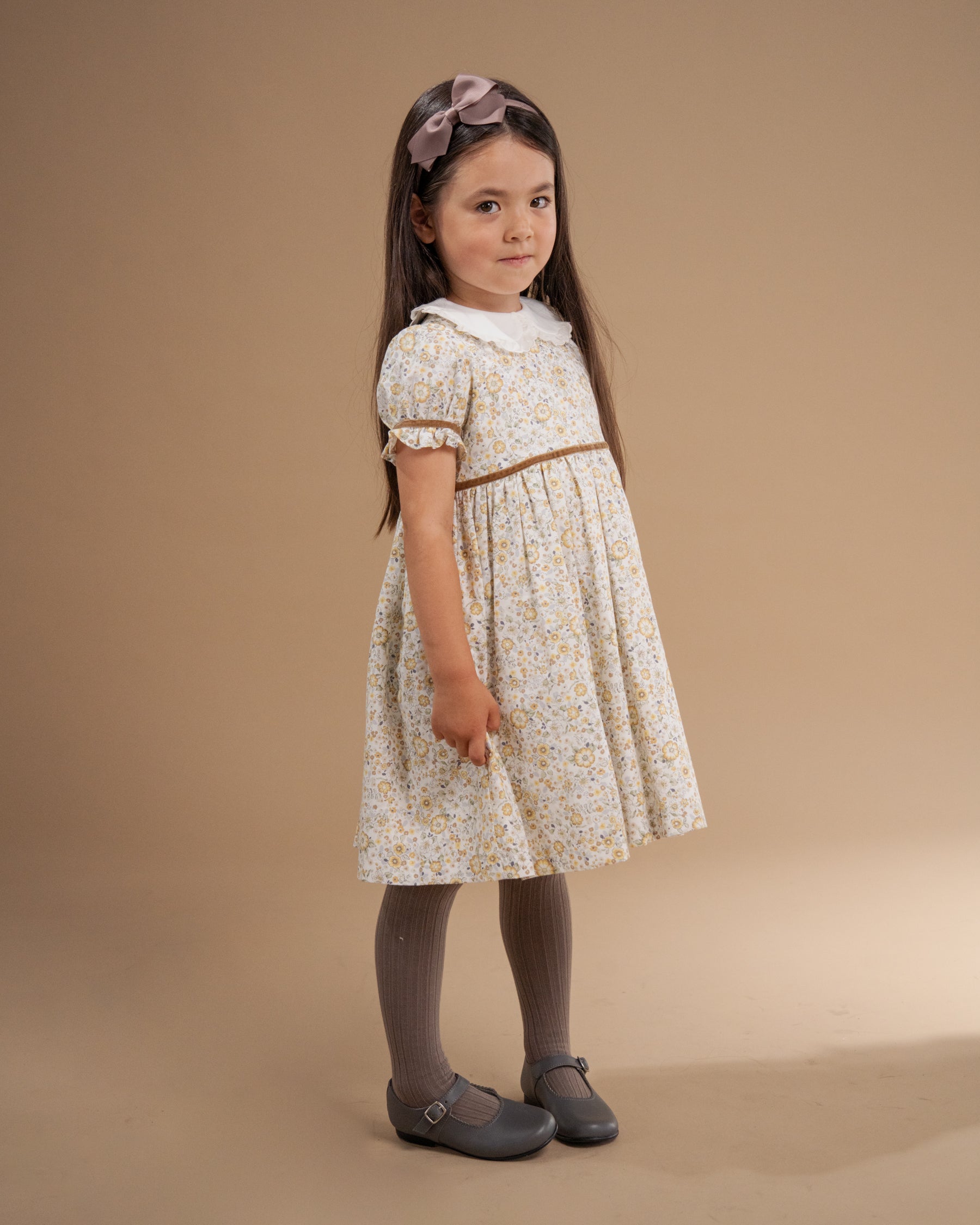 A young girl stands in a studio wearing a short-sleeve cream dress with a small yellow and green floral print, a white collar, and a thin brown waistband. She is also wearing gray tights and gray Mary Jane shoes against a solid warm beige background.