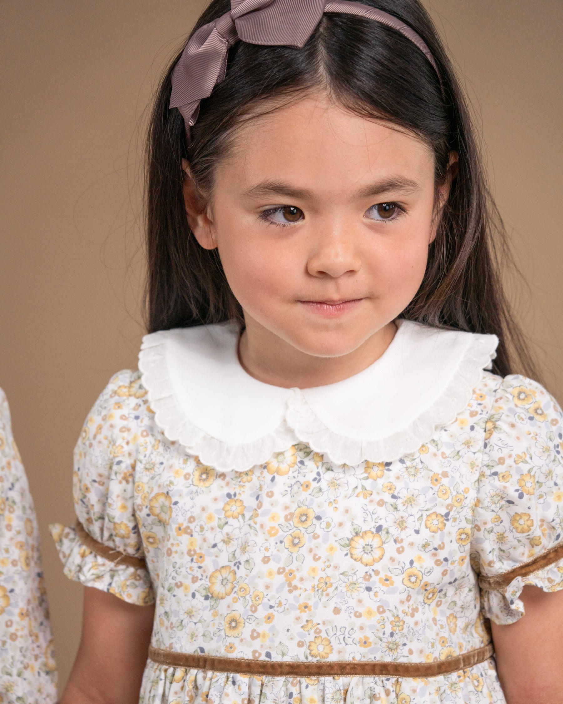 A young girl with dark hair wearing a dusty mauve bow headband and a white floral dress with a Peter Pan collar, photographed against a solid beige background.