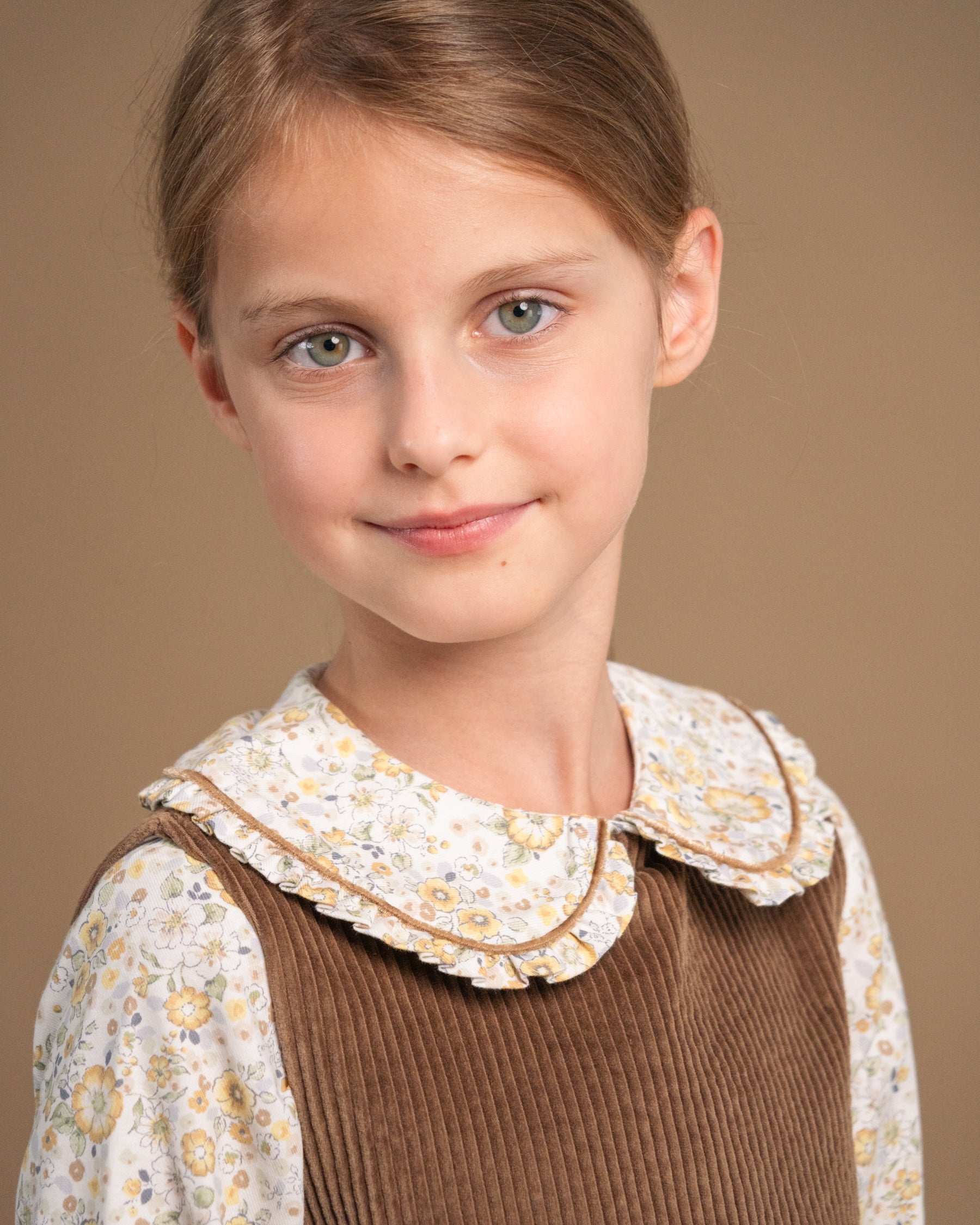 A young girl with light brown hair and green eyes wearing a brown corduroy pinafore vest over a cream floral long-sleeve shirt with a ruffled Peter Pan collar, set against a solid warm brown background.