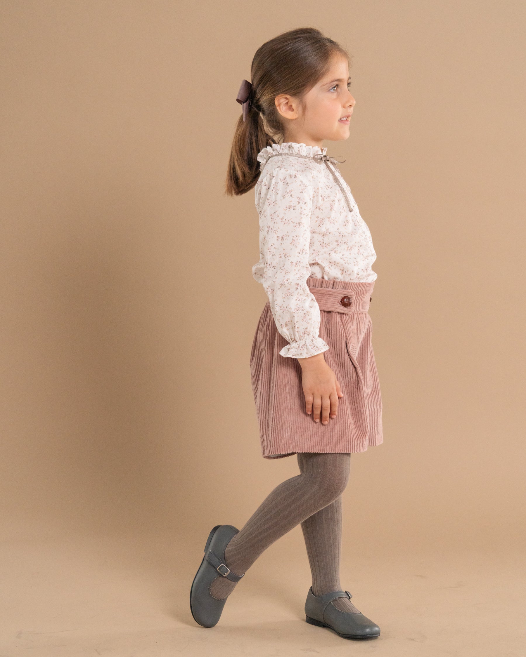 A young girl in profile wearing a white floral long-sleeve blouse, a dusty pink corduroy skirt with a button waist, gray ribbed tights, and gray Mary Jane shoes, set against a solid beige studio background.
