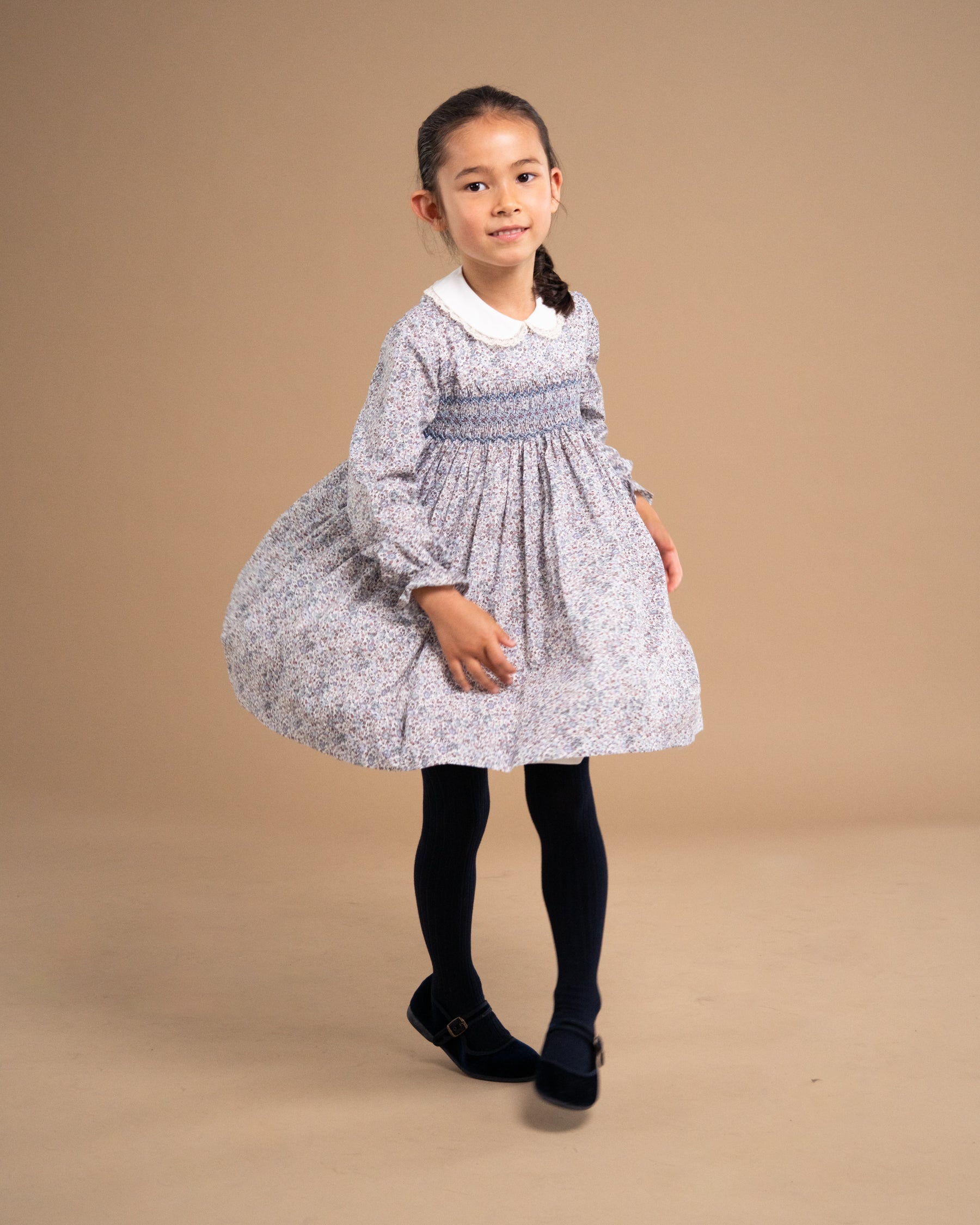 A young girl modeling a light gray and white floral smocked dress with a white Peter Pan collar, black tights, and black shoes, standing against a solid beige studio background.