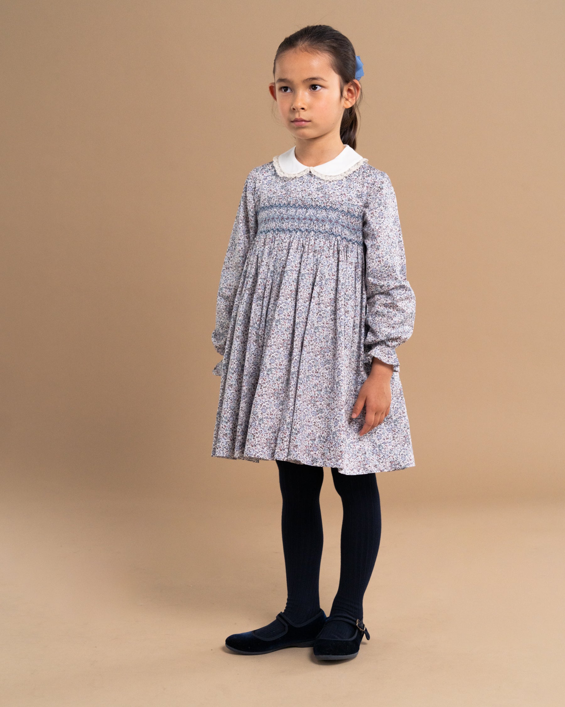 A young girl modeling a long-sleeve smocked dress with a blue and white floral pattern, a white Peter Pan collar, black tights, and black shoes, standing against a solid beige studio background.