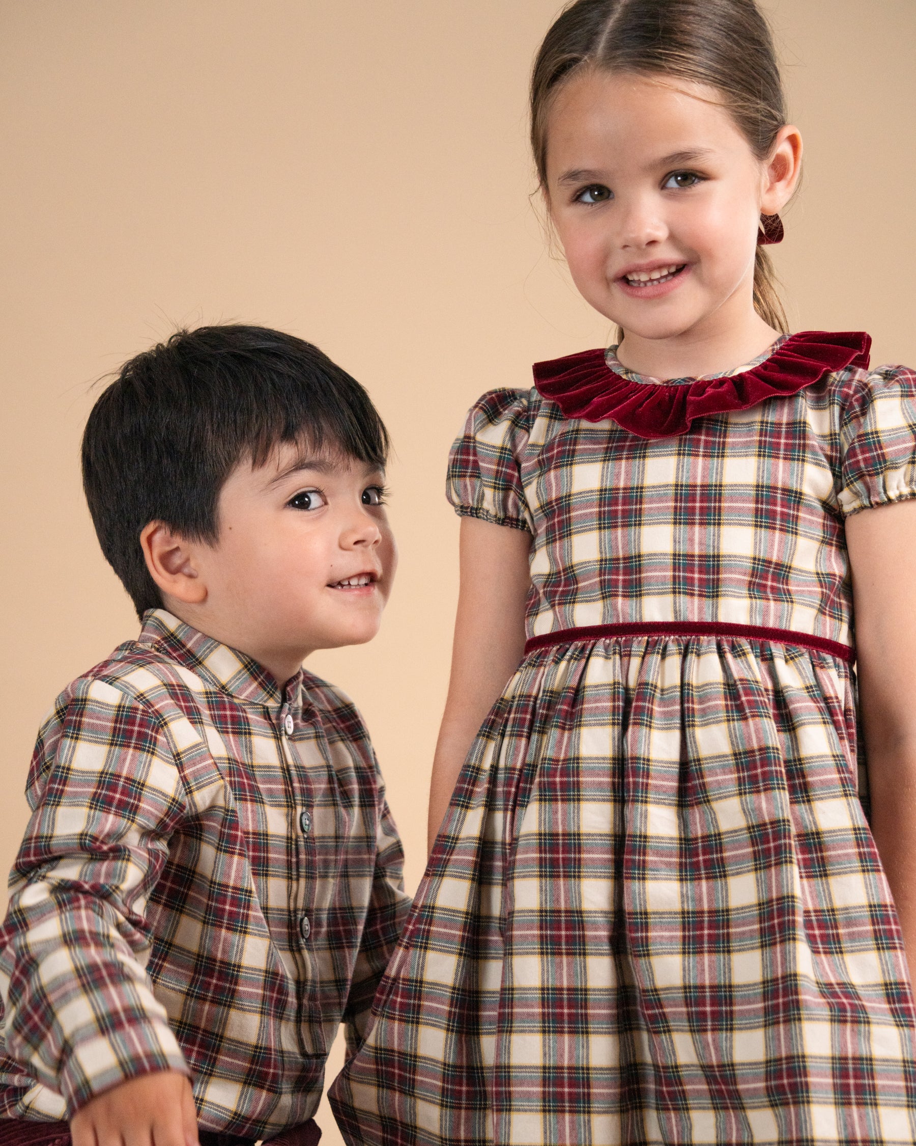 A young boy and girl modeling matching plaid children's outfits against a solid beige studio background. The boy wears a matching plaid long-sleeve button-up shirt, and the girl wears a matching plaid dress with a dark red ruffled collar and waistband.