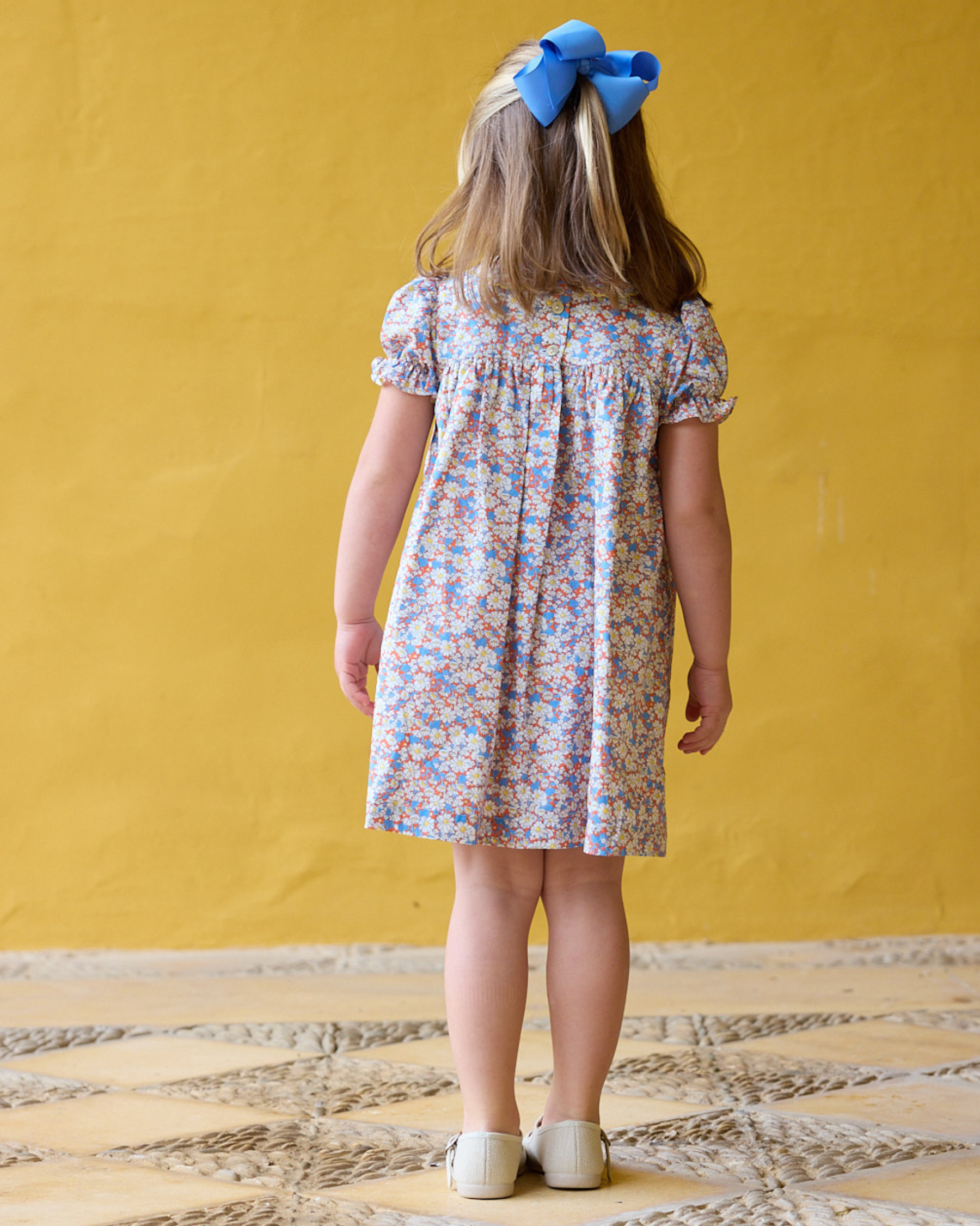 Young girl wearing a floral dress with puffed sleeves against a yellow wall.