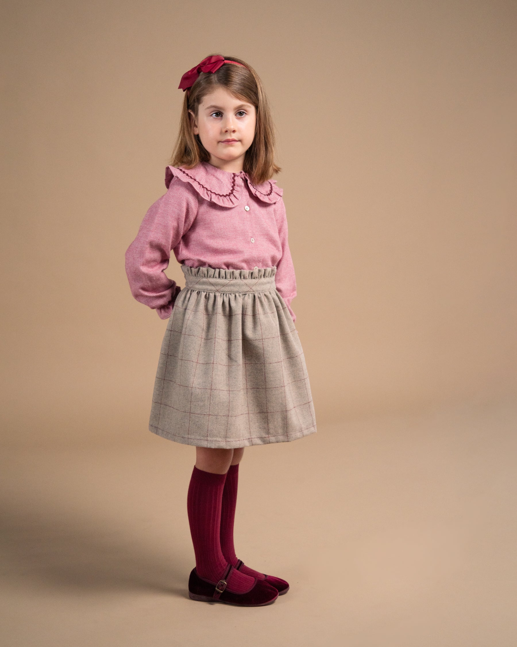 A young girl stands against a solid beige studio background wearing a pink long-sleeve blouse with a ruffled collar, a pleated light gray plaid skirt, dark red knee-high socks, and dark red shoes, with a matching red bow in her hair.