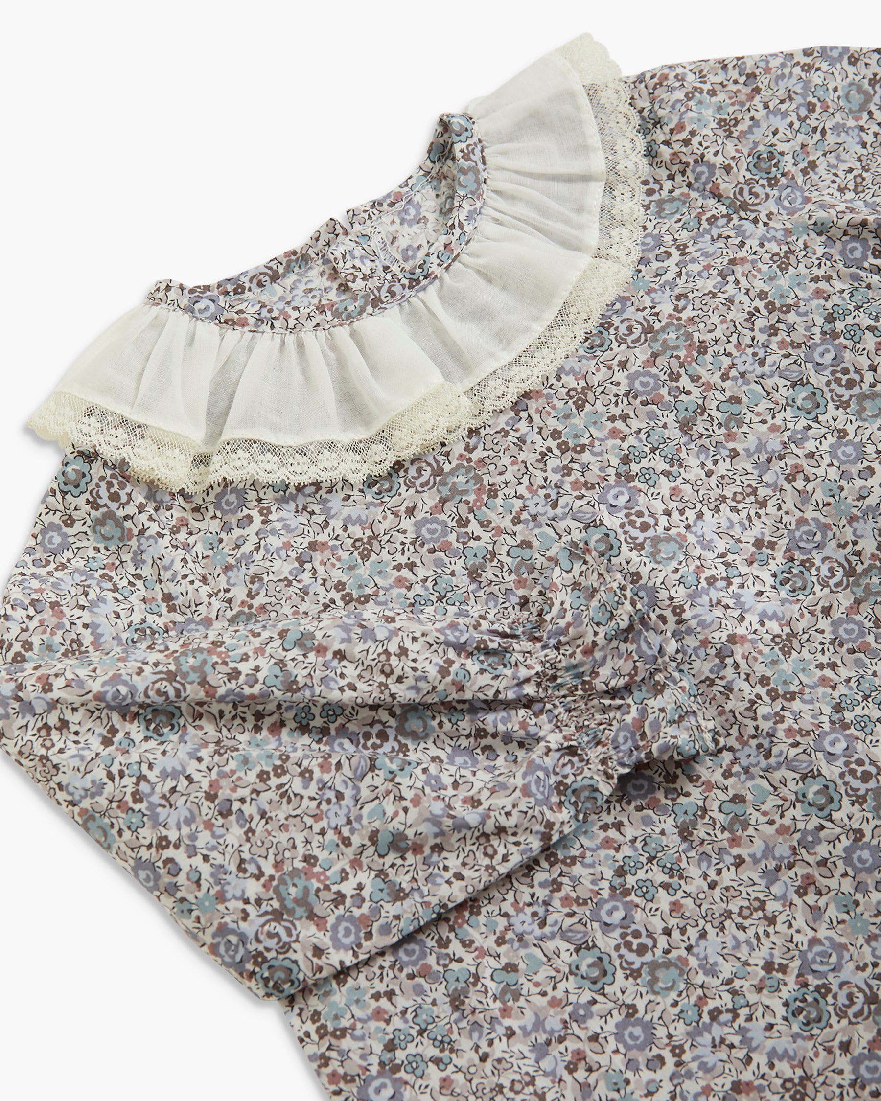A close-up of a long-sleeve blouse featuring a dense floral and paisley print in muted blue, brown, and beige tones, with a prominent ruffled collar trimmed in cream lace.