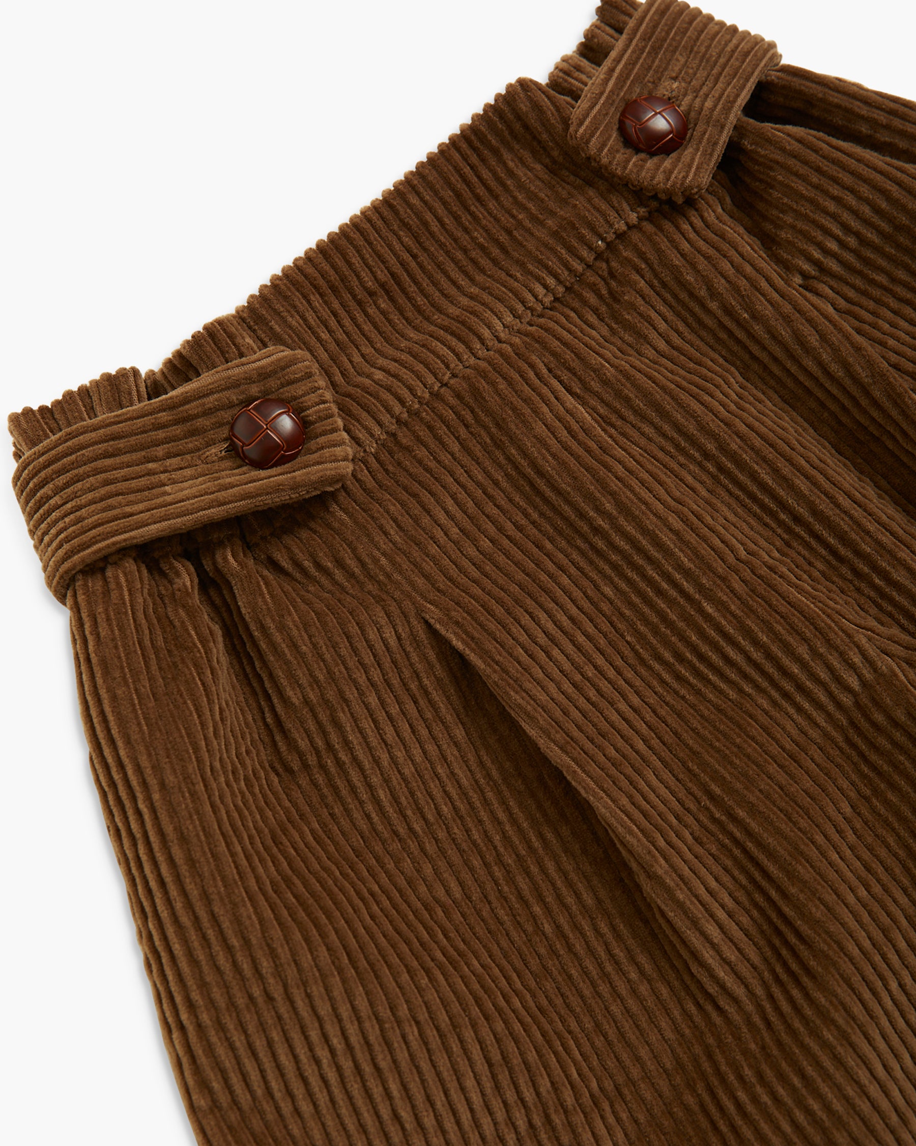 Close-up of brown corduroy trousers showing the pleated fabric texture and buttoned waistband details against a white background.