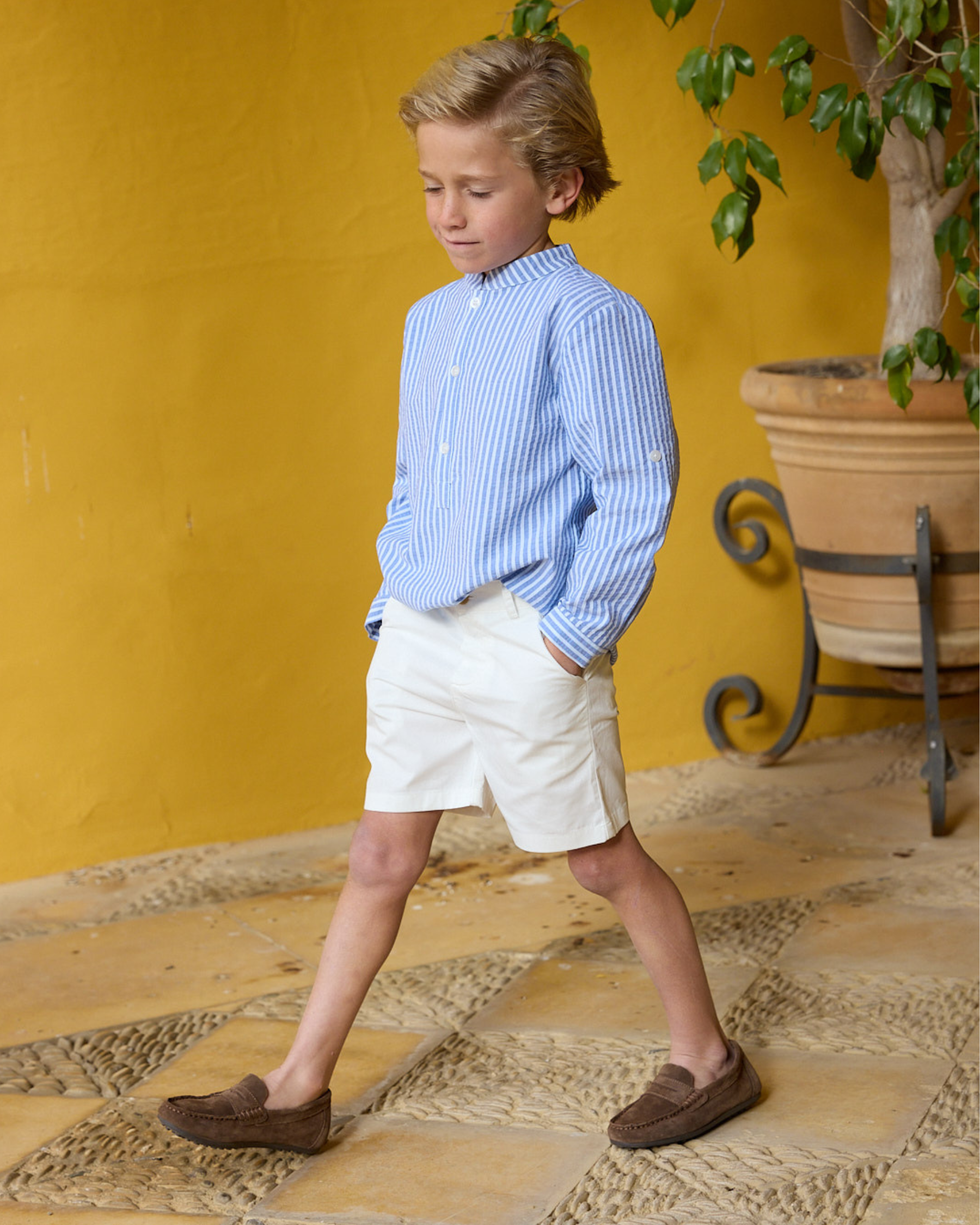Young boy in a blue striped shirt and white shorts standing in front of a yellow wall with a plant.