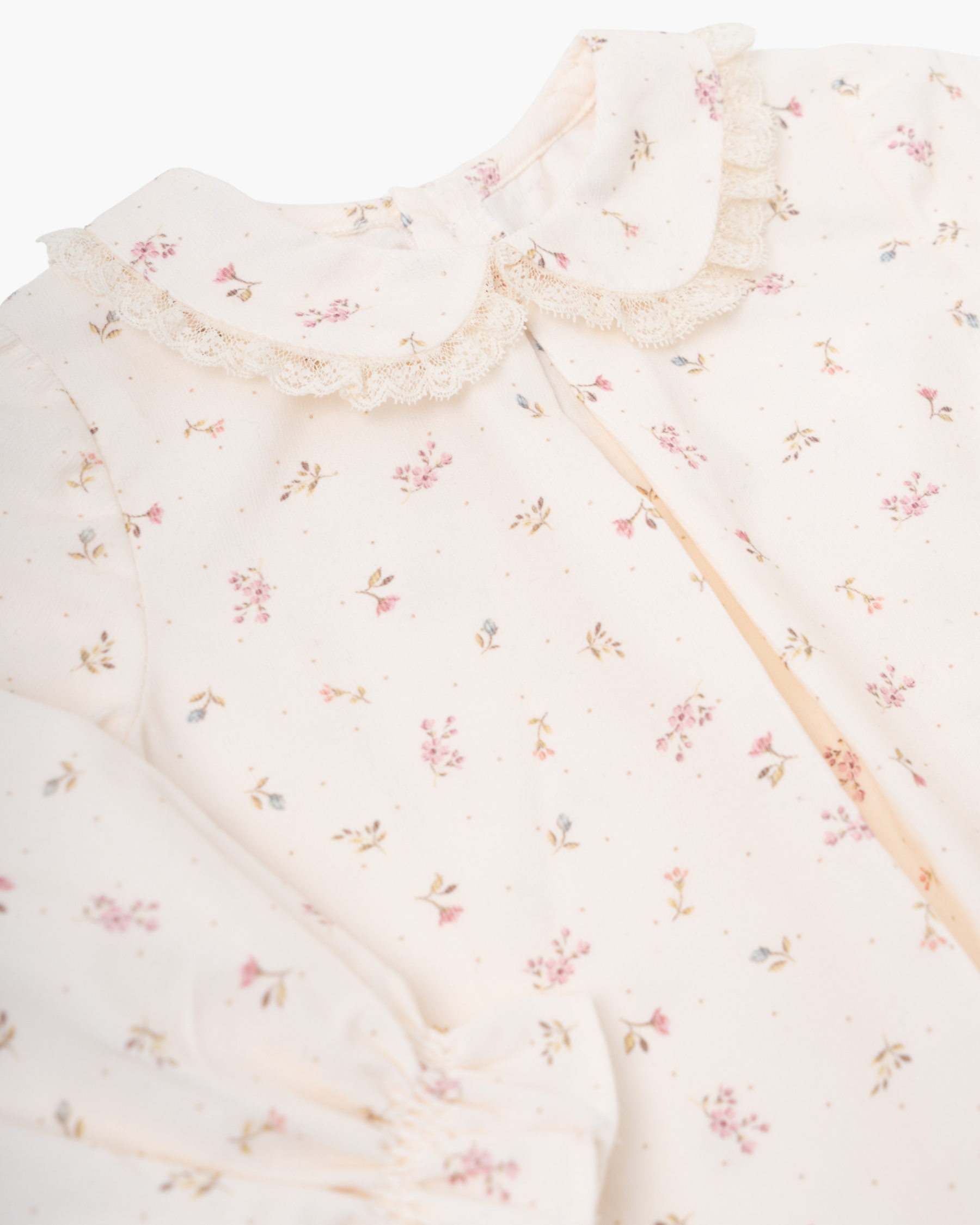 A close-up of a cream-colored baby dress or long-sleeve top featuring a delicate pink and green floral print, a lace Peter Pan collar, and a soft gathered hem.