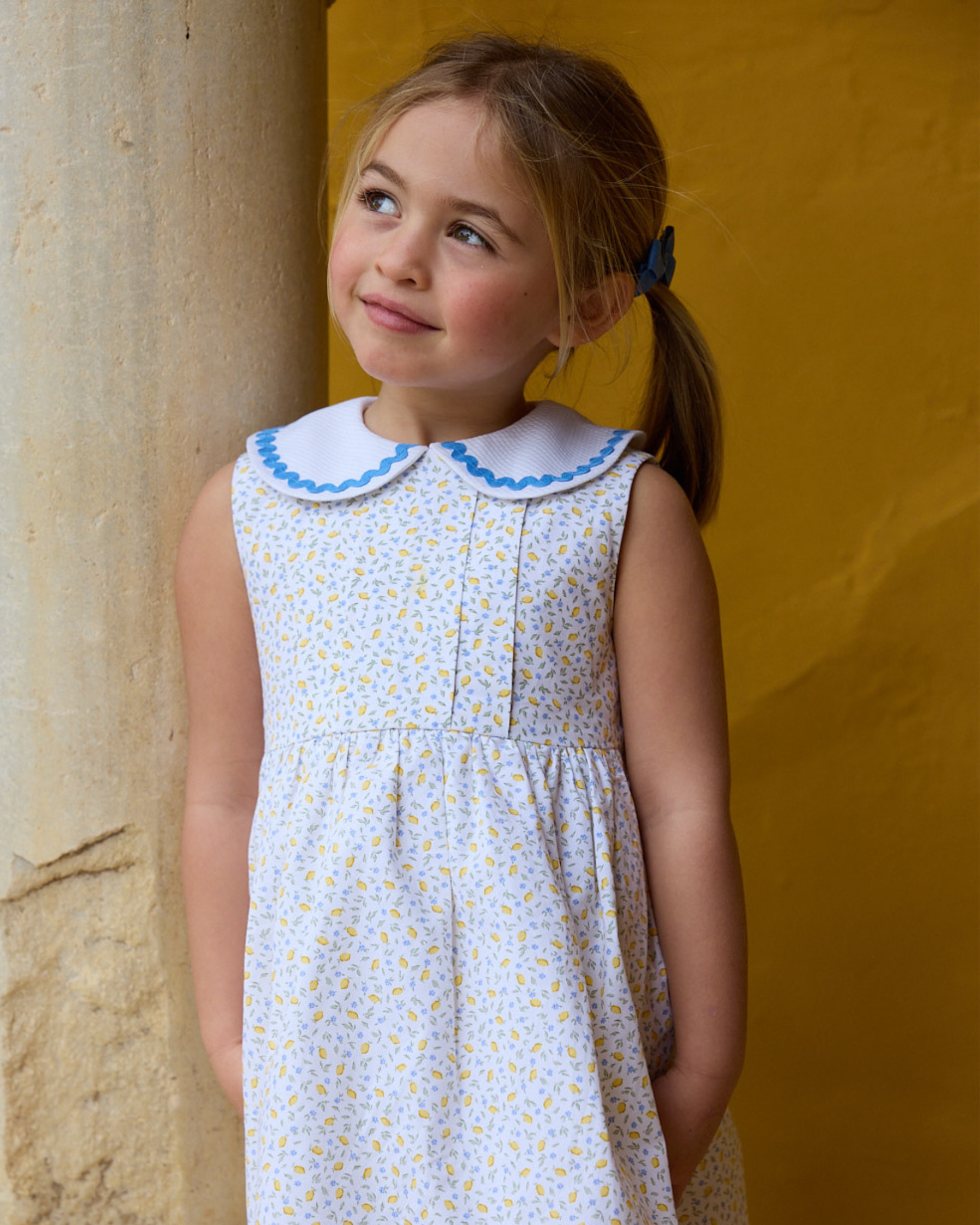 Young girl wearing a white dress with blue collar against a yellow wall.