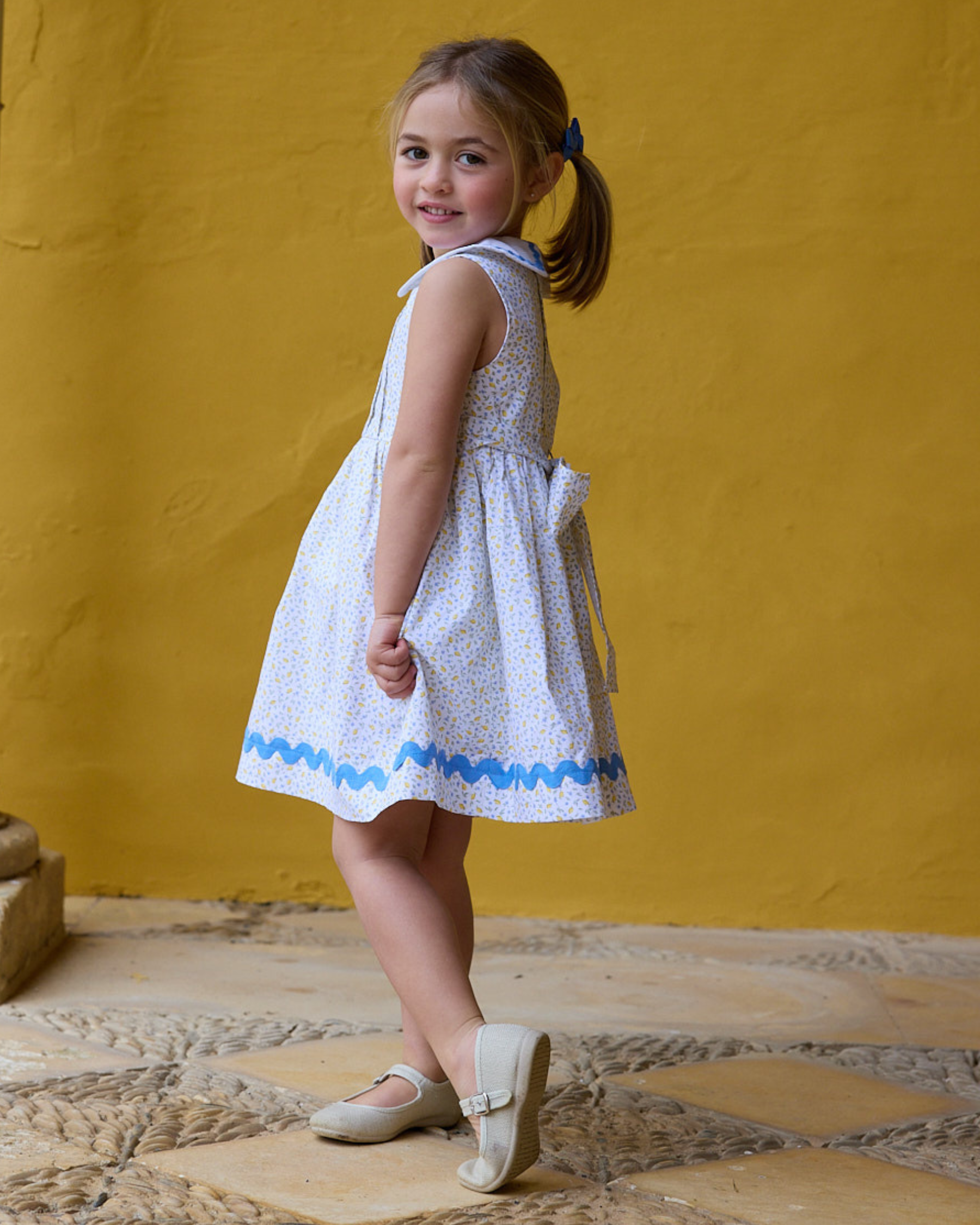 Young girl in a white dress with blue trim standing against a yellow wall.