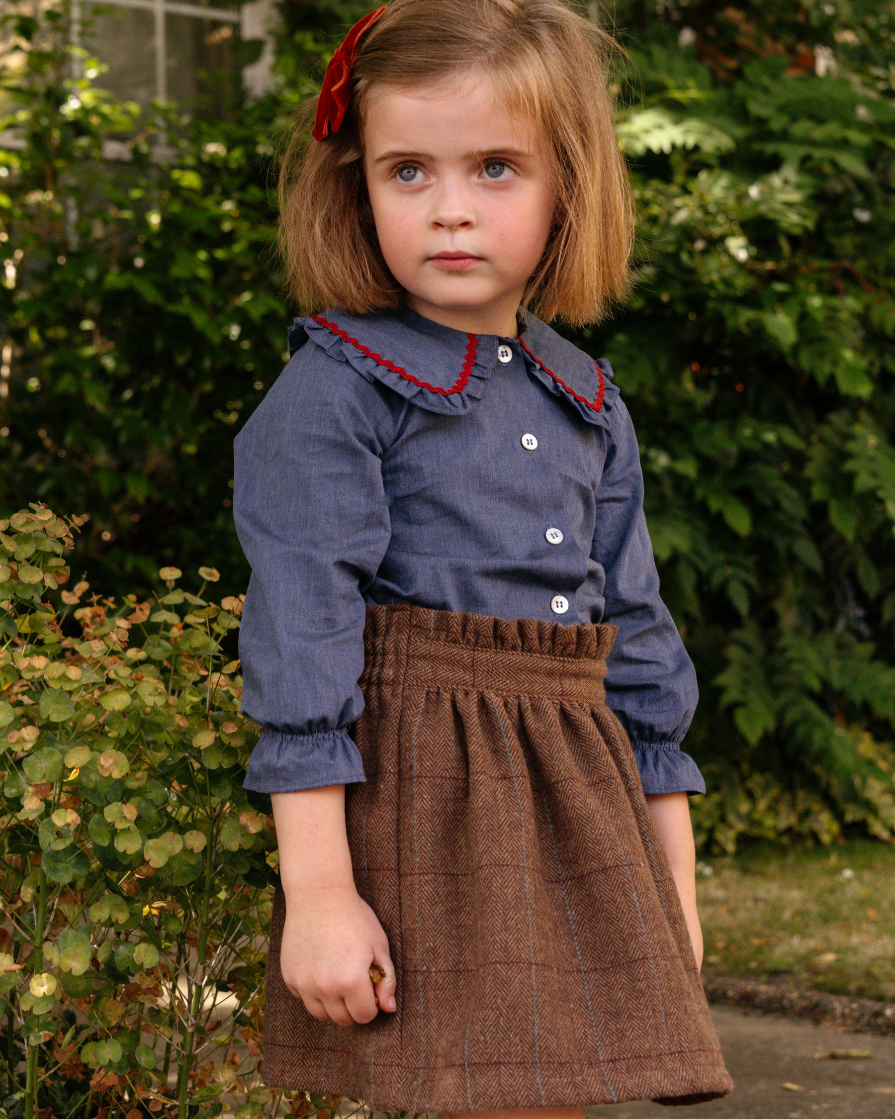 Young girl wearing a blue shirt with red trim and a brown skirt outdoors.