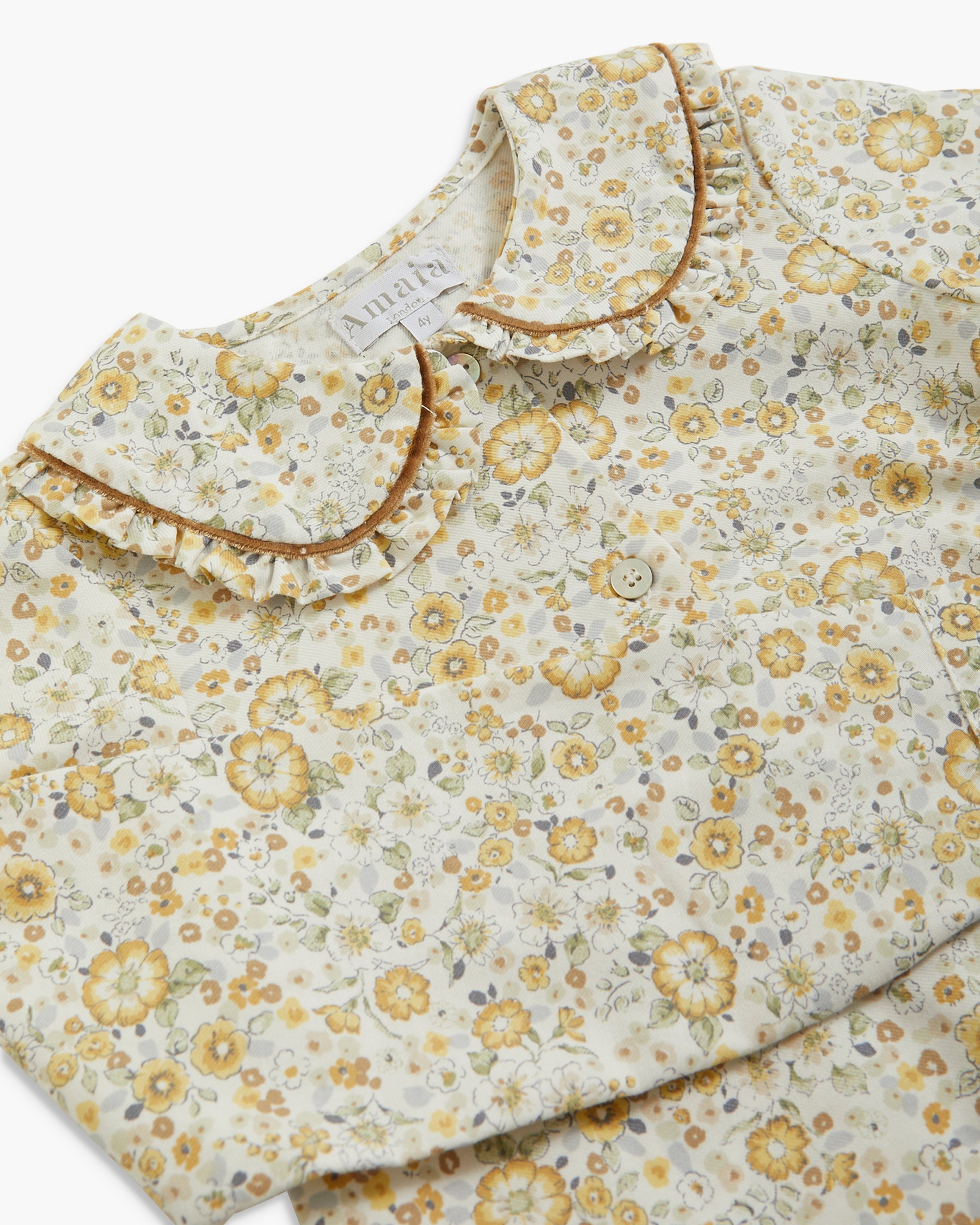 A close-up of a children's blouse with a delicate yellow and green floral print on a cream background, featuring a ruffled Peter Pan collar and a small smiley face button.