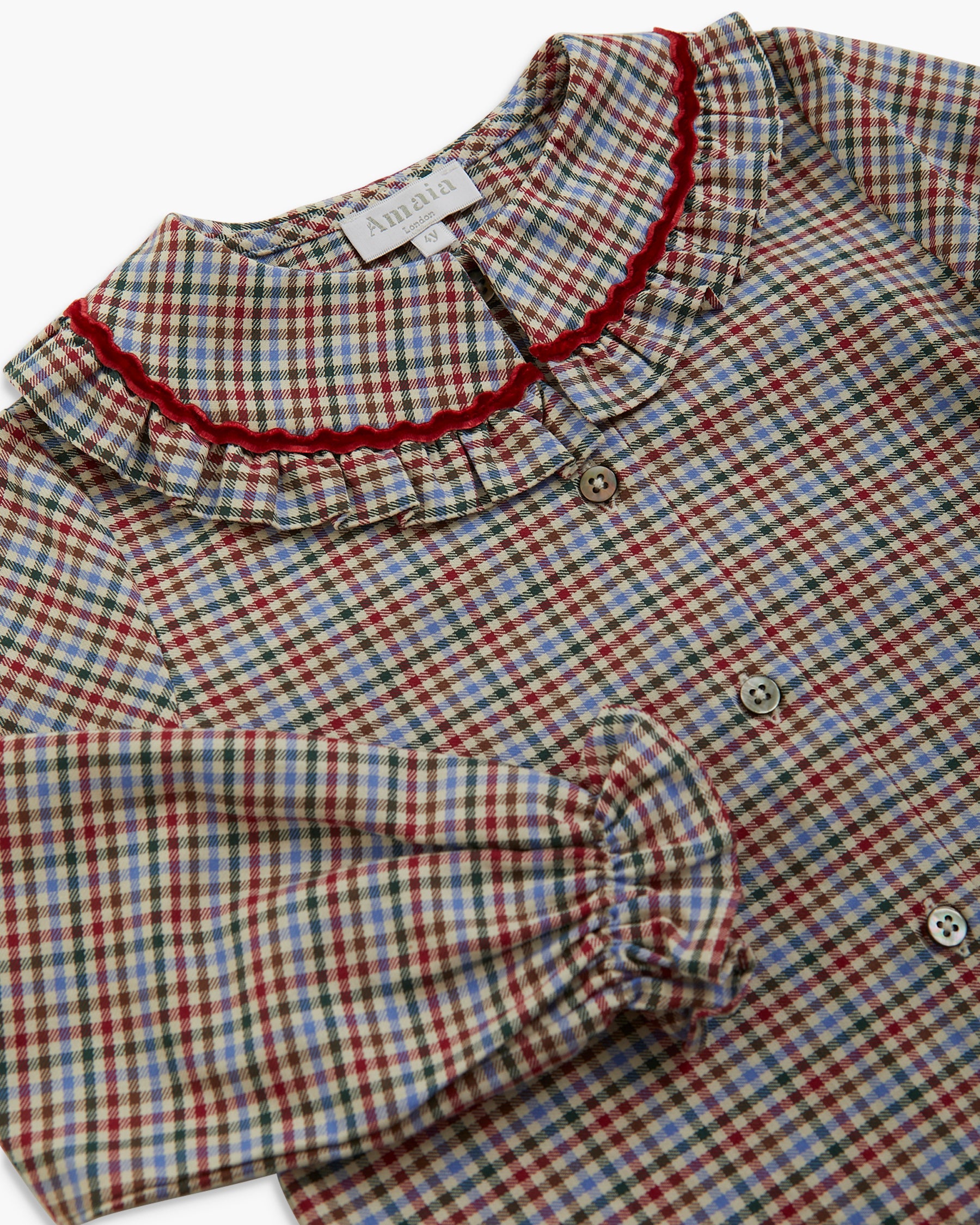 A close-up of a children's long-sleeve blouse with a red, blue, and beige gingham pattern, featuring a ruffled collar trimmed with red embroidery and a gathered elbow-length sleeve.