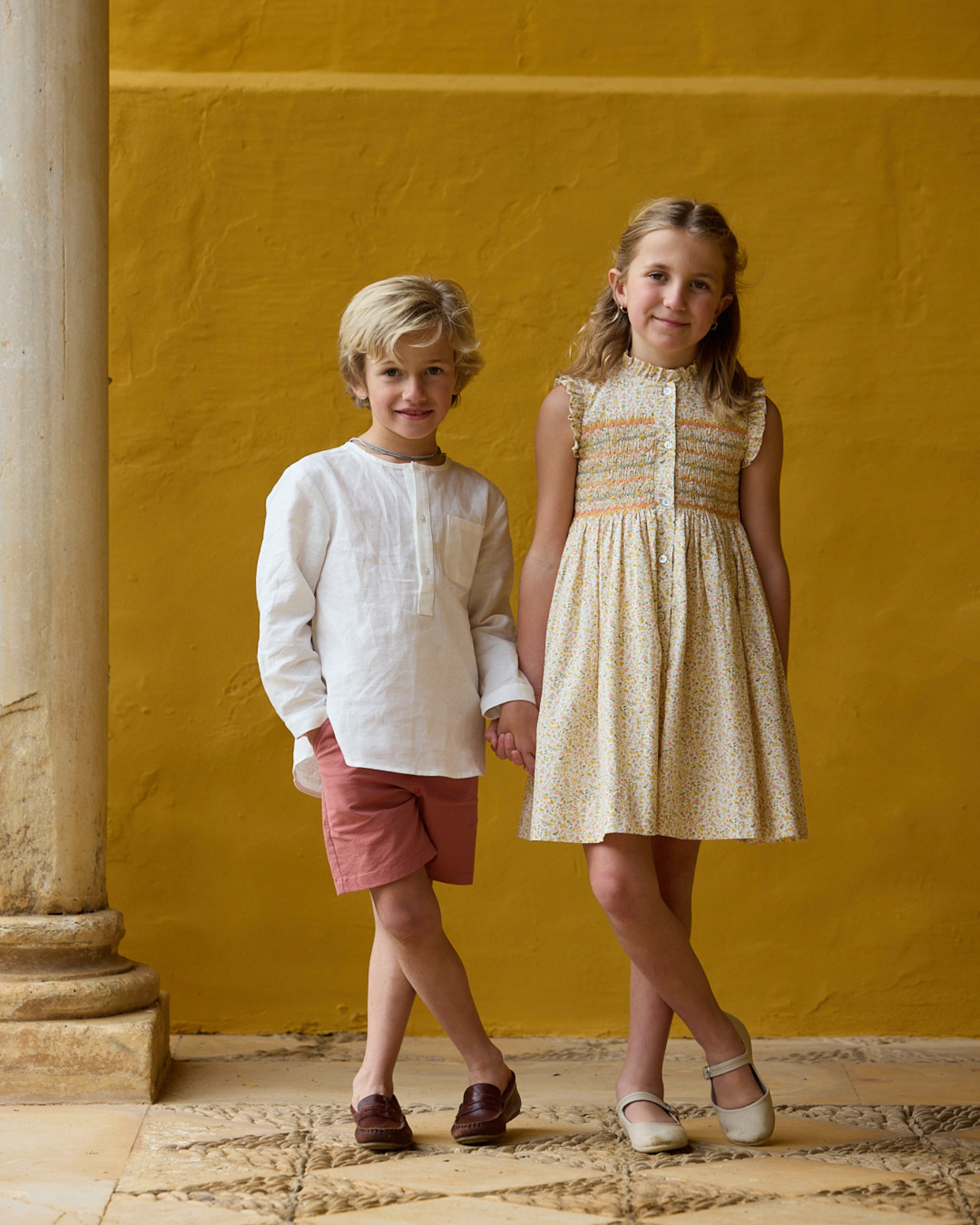 Two children standing against a yellow wall with a stone column.