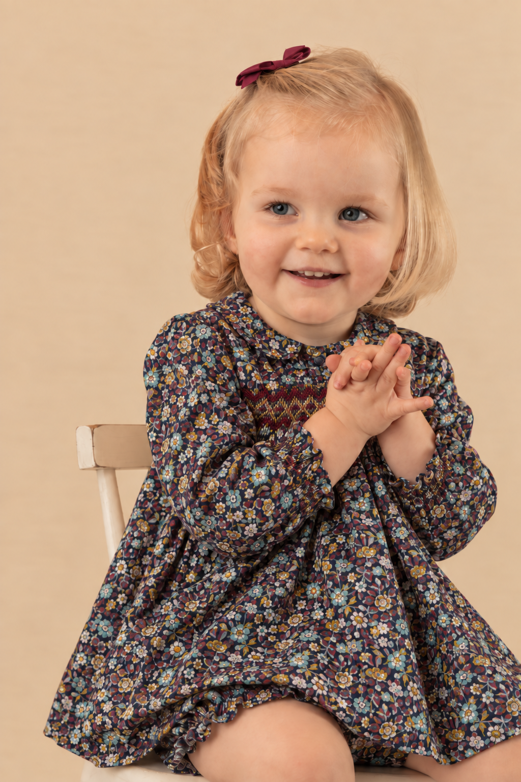 A young toddler girl with blonde hair and a red bow sits on a small wooden chair, wearing a dark floral long-sleeve dress with a patterned yoke. The background is a solid warm beige studio backdrop.