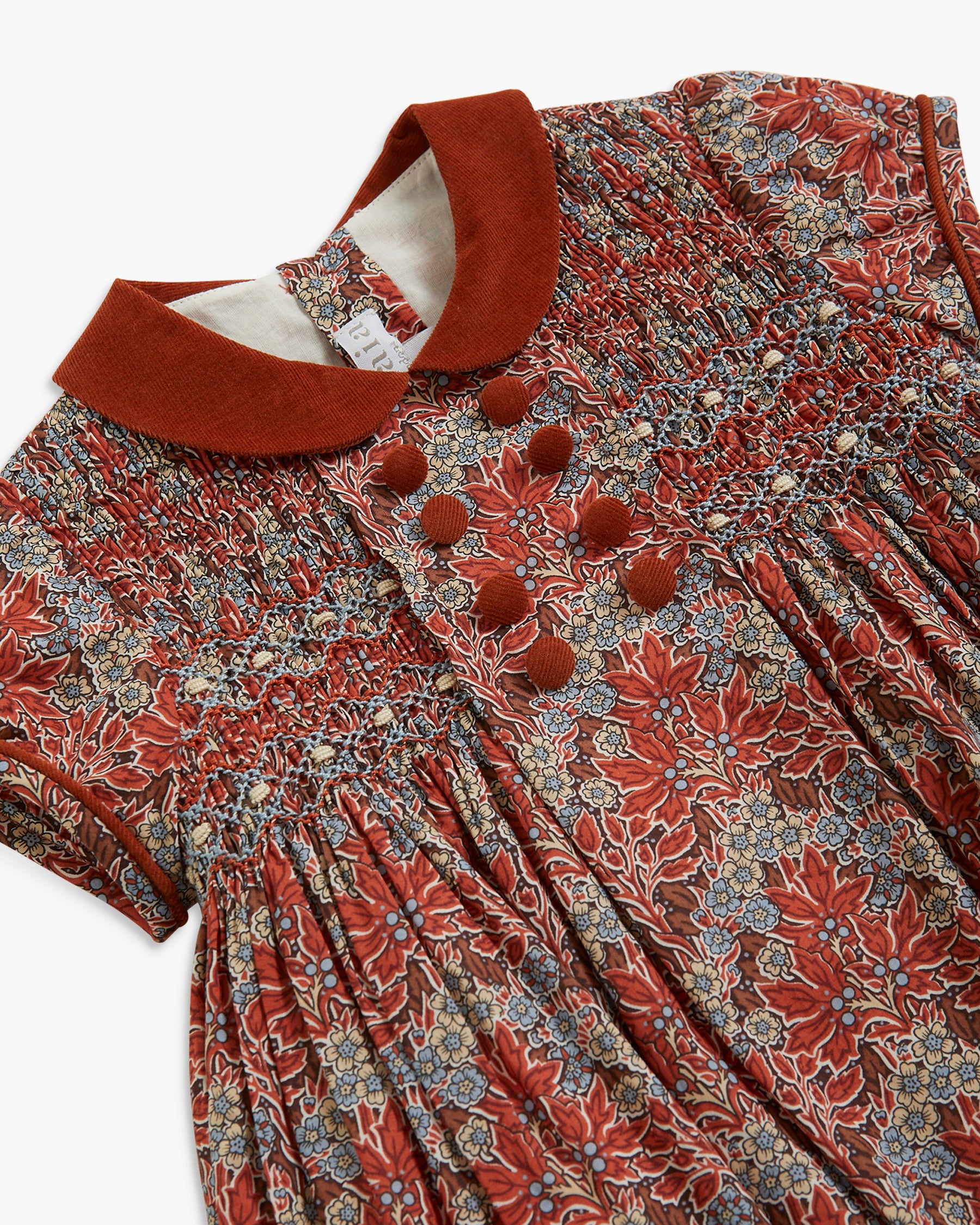 A close-up of a short-sleeve dress with a smocked bodice, featuring a rust red collar and matching decorative buttons, over a dense floral and botanical print in rust red, beige, and blue.
