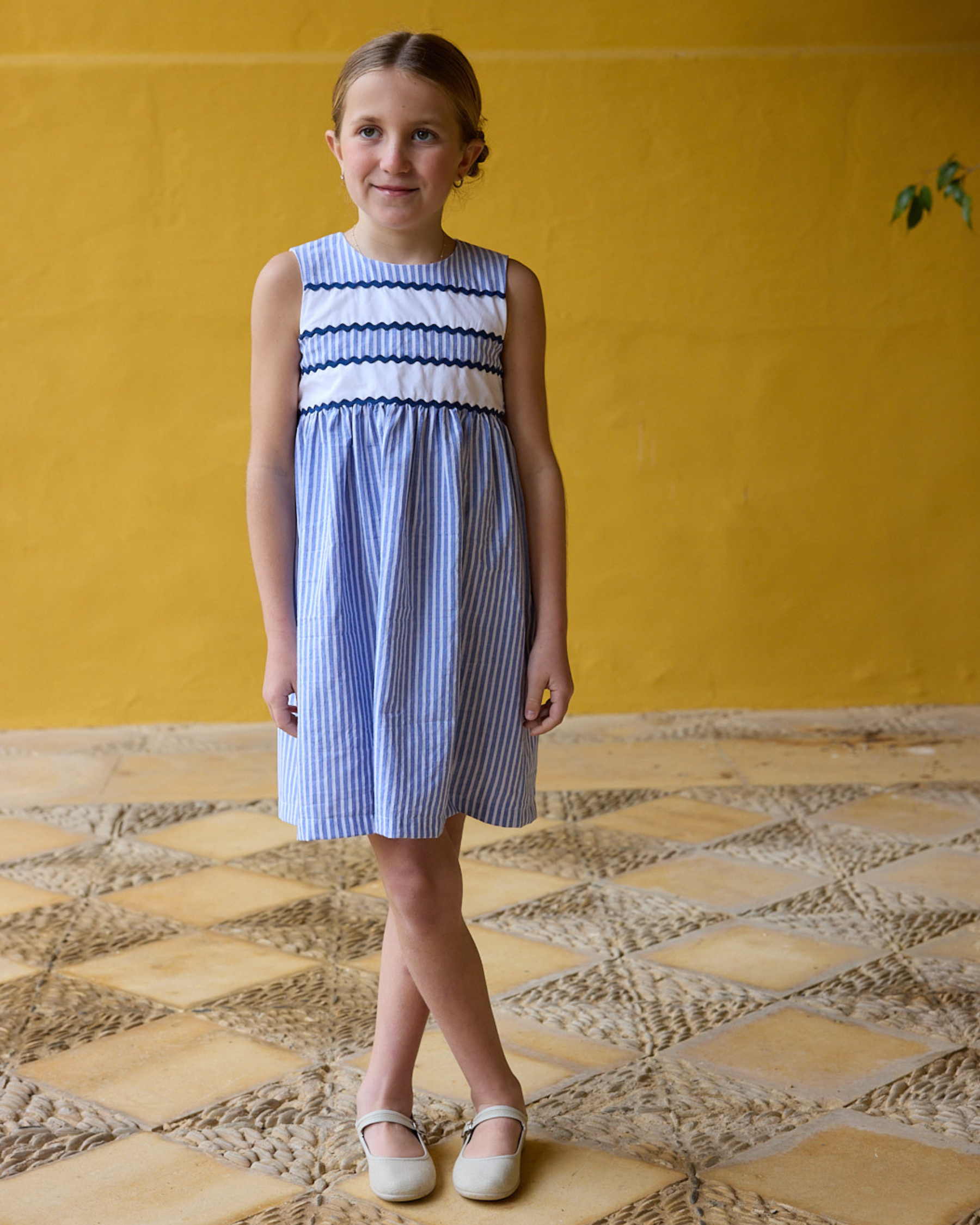 Young girl wearing a blue and white dress standing on a patterned floor with a yellow wall in the background.