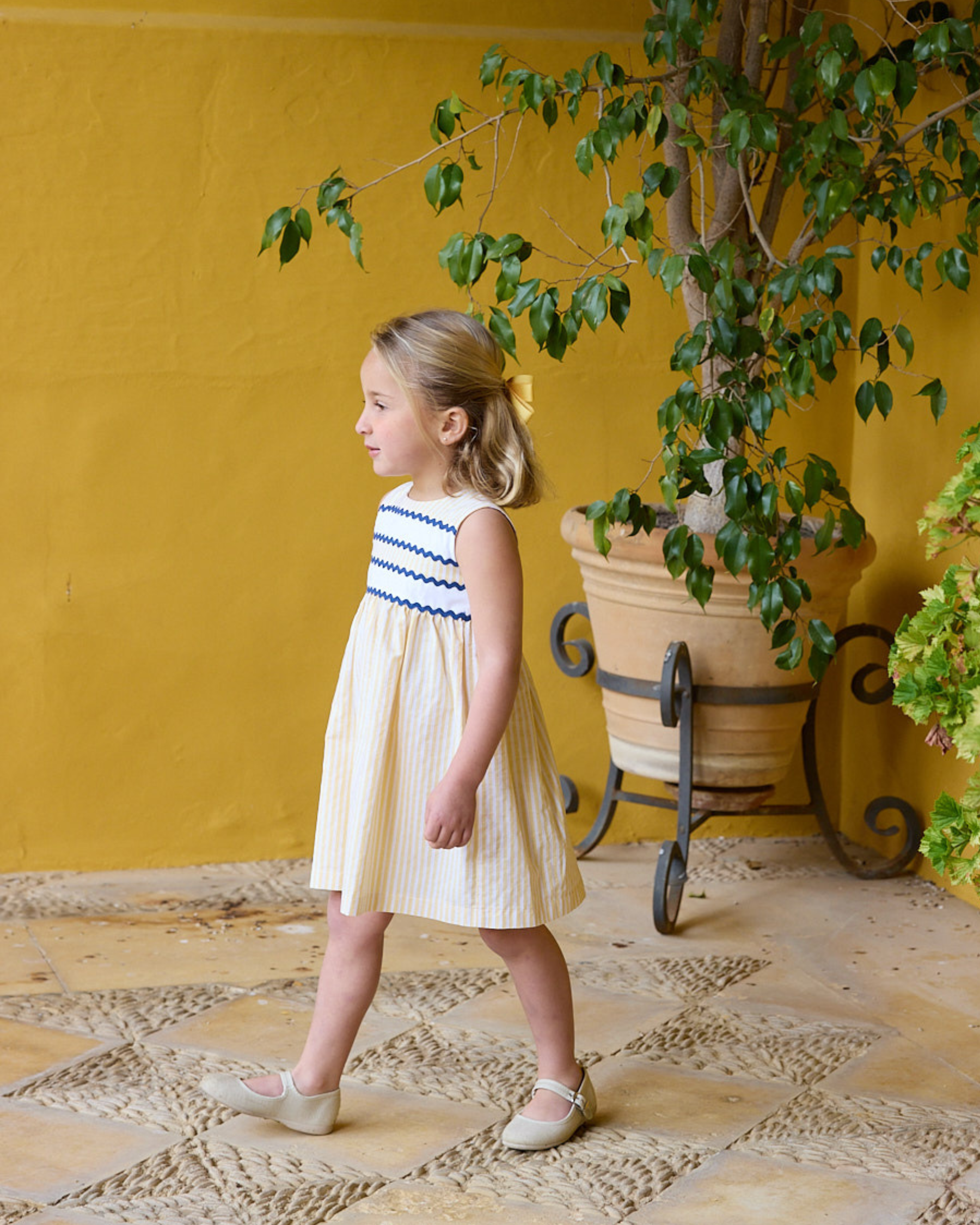 Young girl in a yellow dress standing in front of a yellow wall with a plant.